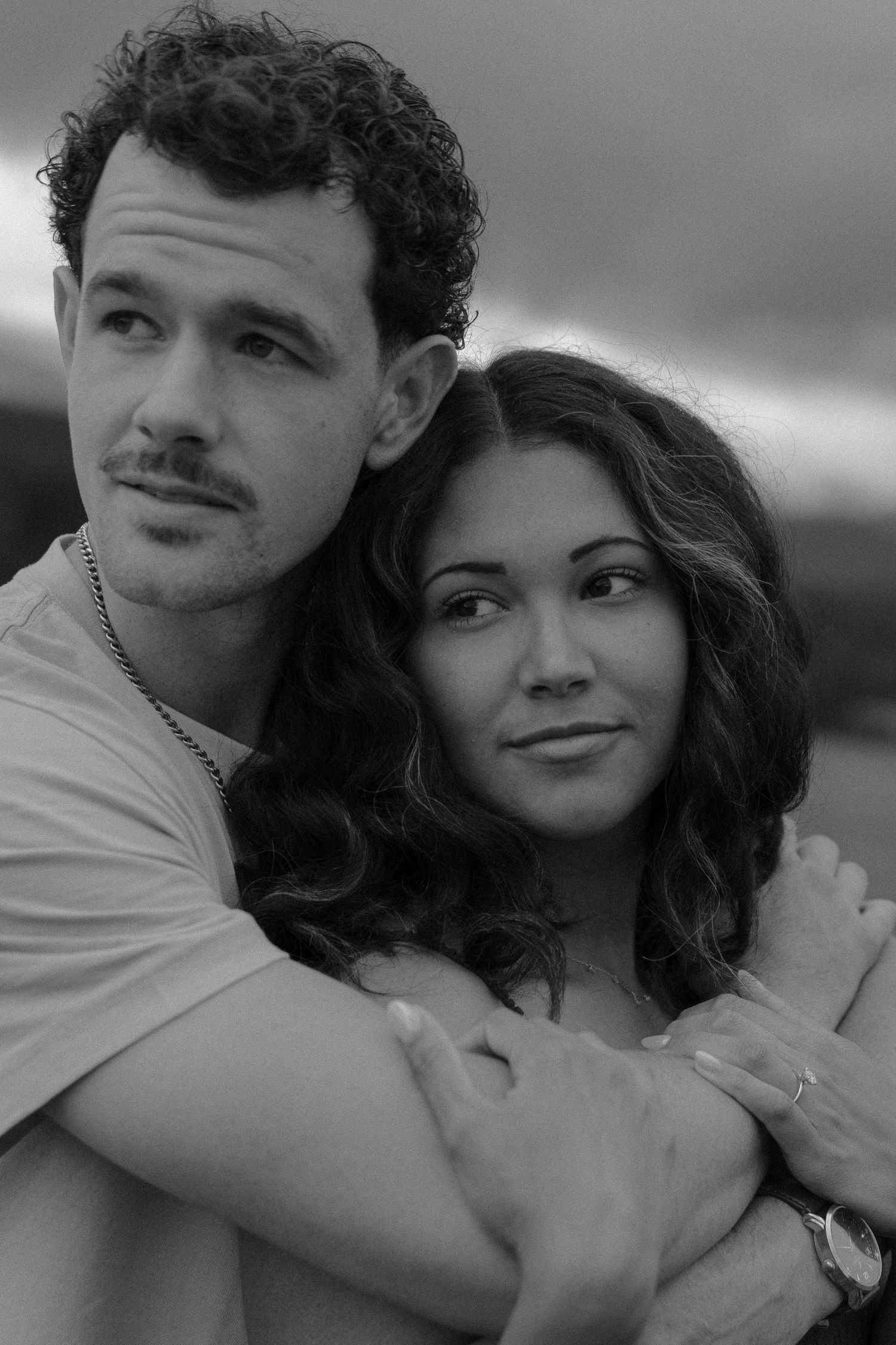 Black and white portrait of a couple embracing outdoors, woman wearing a watch, man in a t-shirt with curly hair.