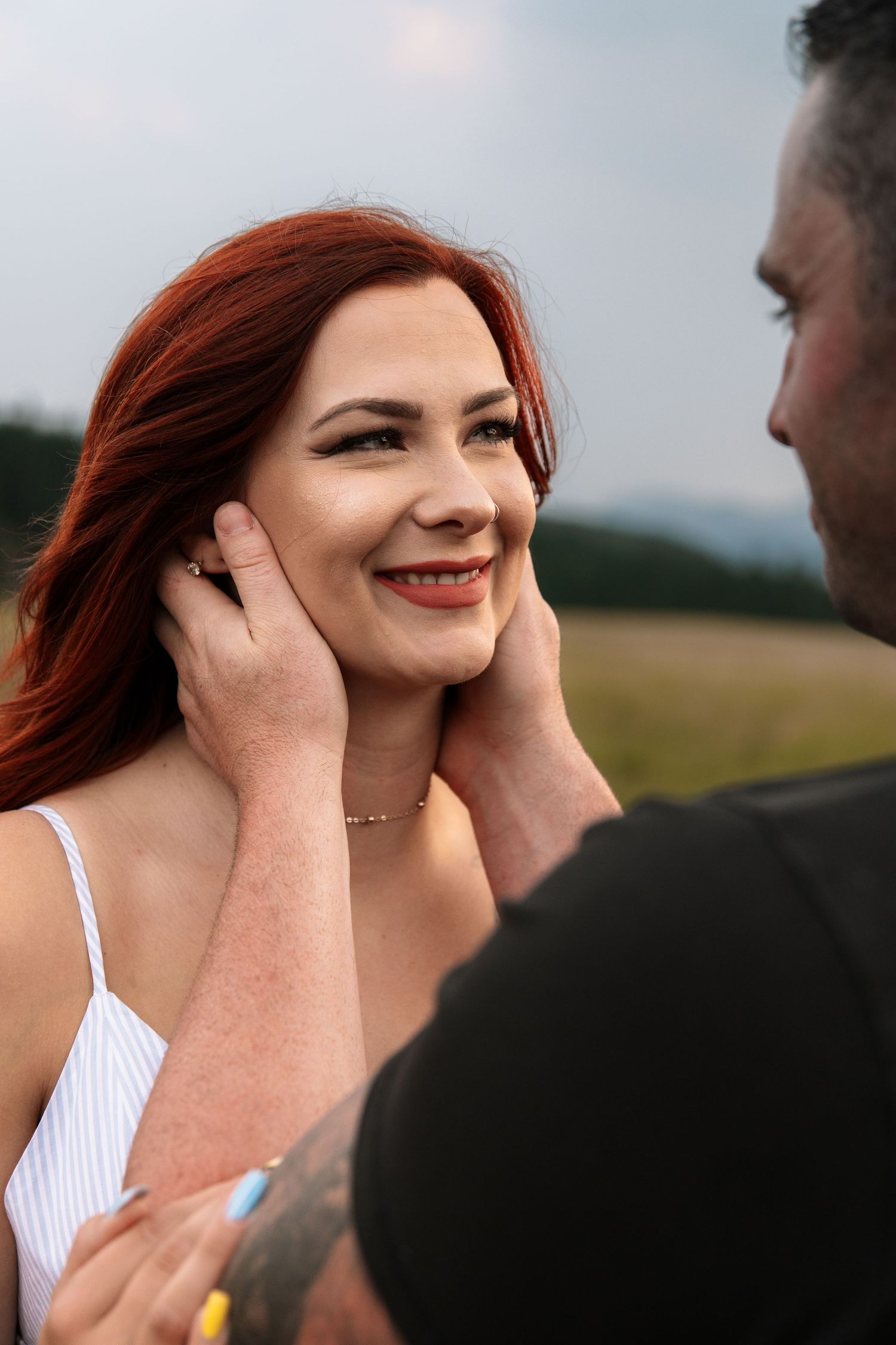 Man gently holding red-haired woman's face as she smiles warmly in an outdoor field setting.