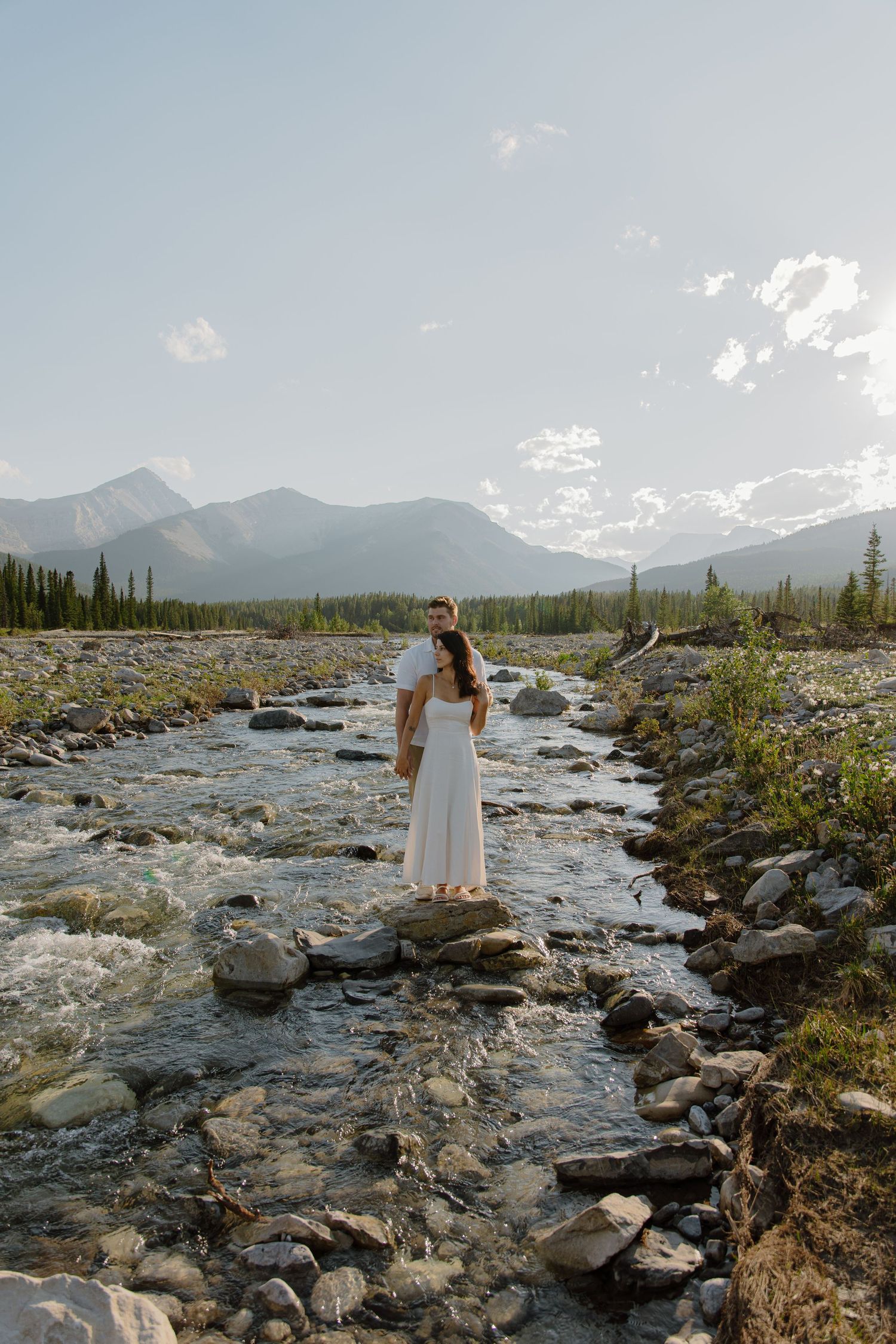 Couple standing on rocks in a mountain stream, woman in white dress, scenic wilderness backdrop with mountains and trees.