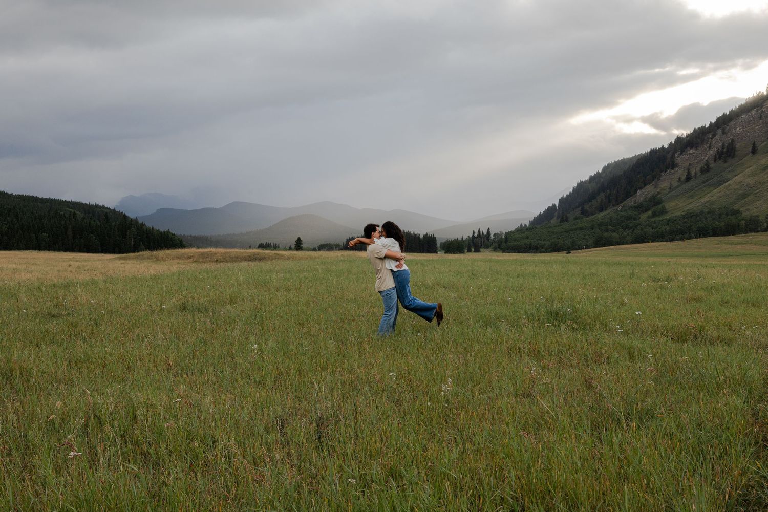 Person dancing joyfully in a vast green meadow with mountains and dramatic storm clouds in the background.
