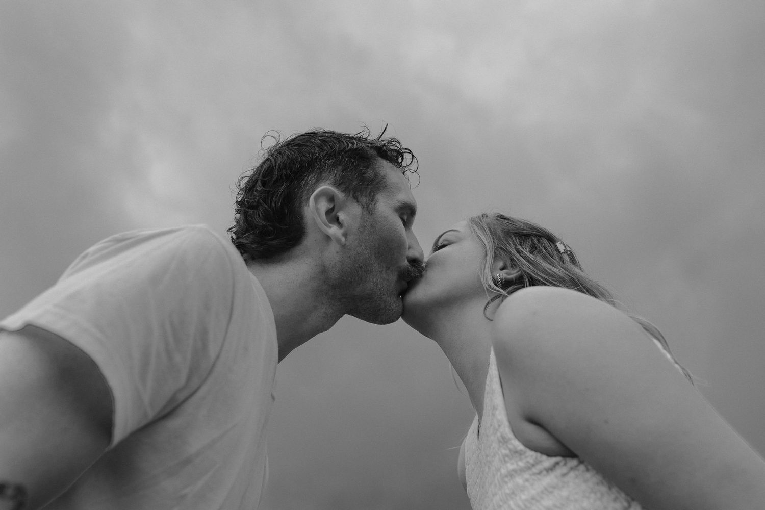 Black and white photo of a couple sharing a romantic kiss against cloudy sky.
