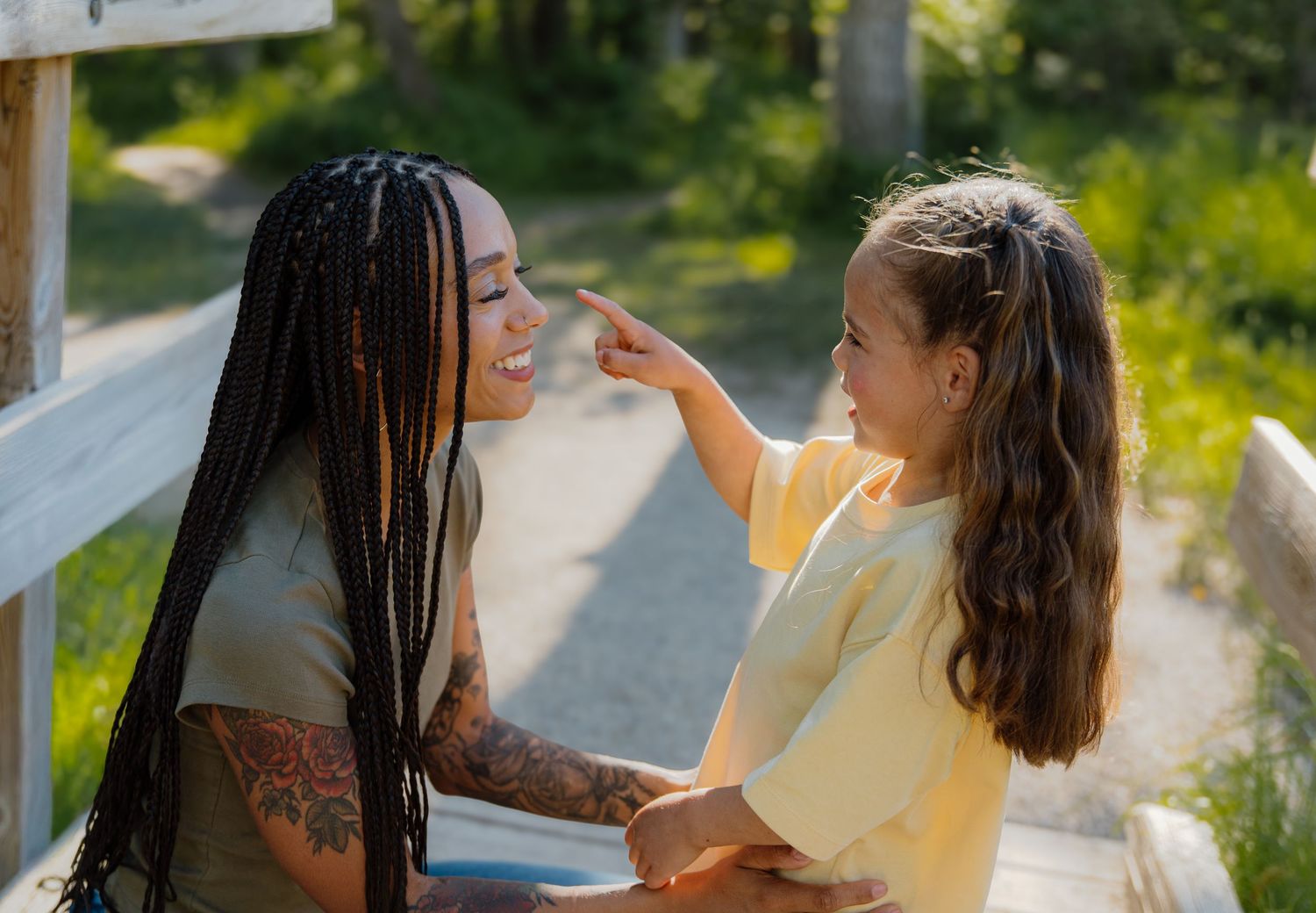 A tattooed woman with braids smiles as a young girl gently touches her face outdoors on a sunny day.