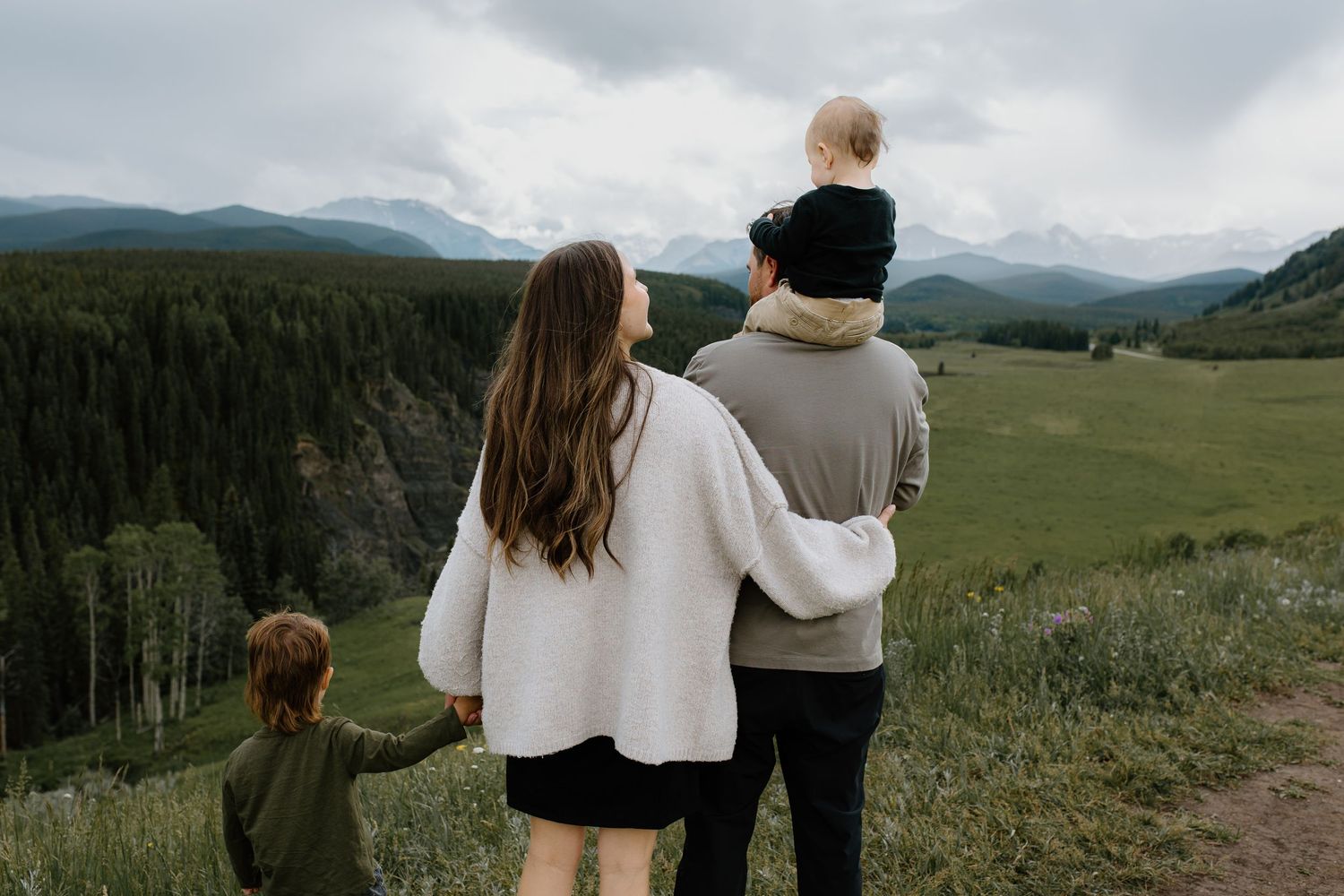 Family of four standing together overlooking a stunning mountain valley landscape from a grassy ridge.