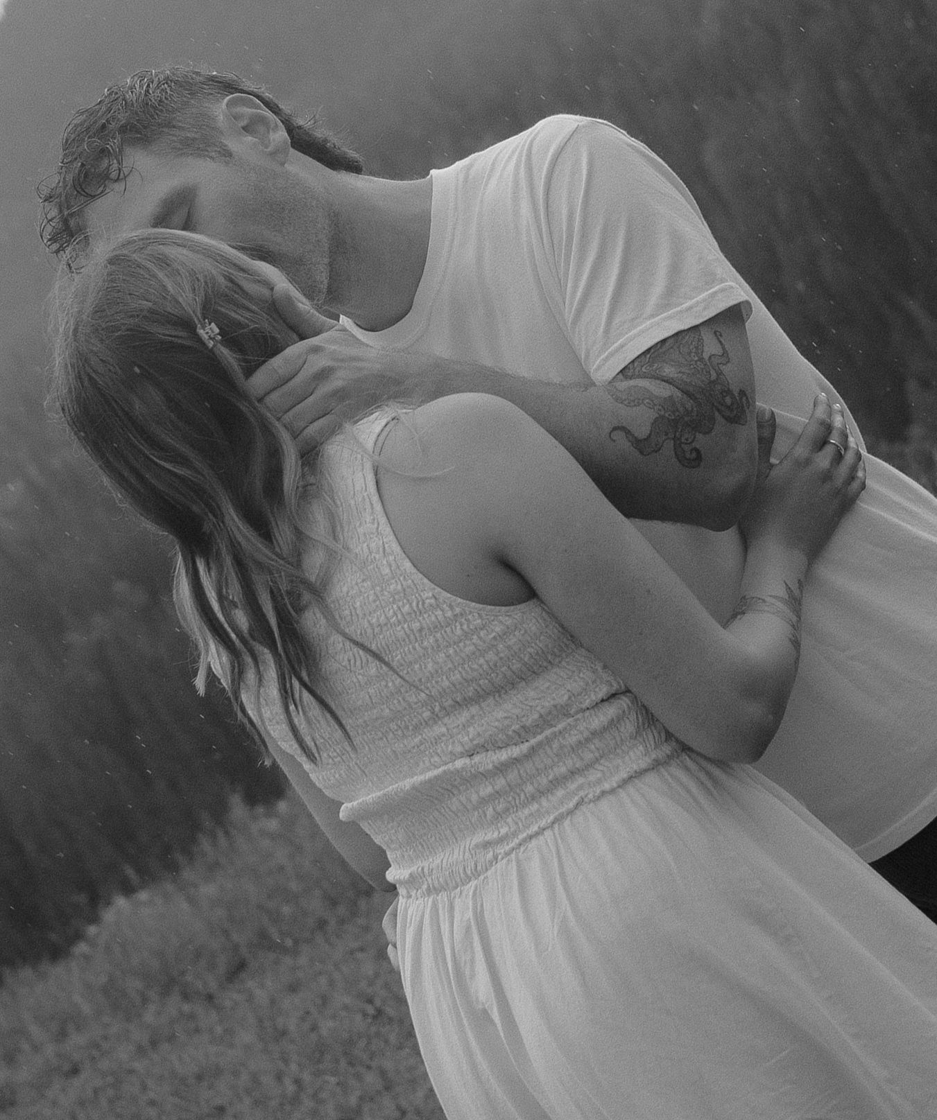 Black and white photo of couple embracing tenderly, man with tattoos kissing womans head.