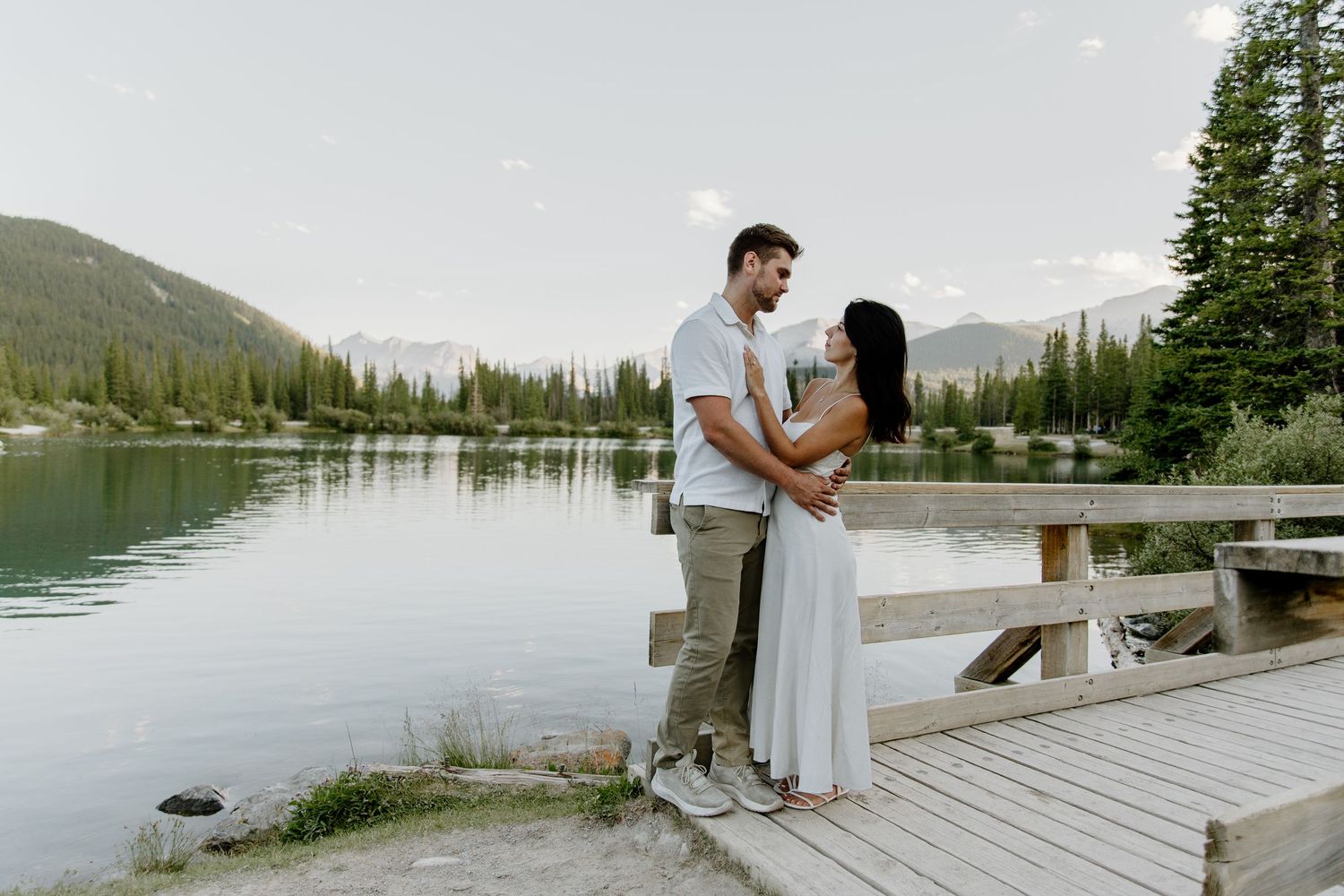 Couple embracing on a wooden dock by a serene mountain lake surrounded by evergreen trees.