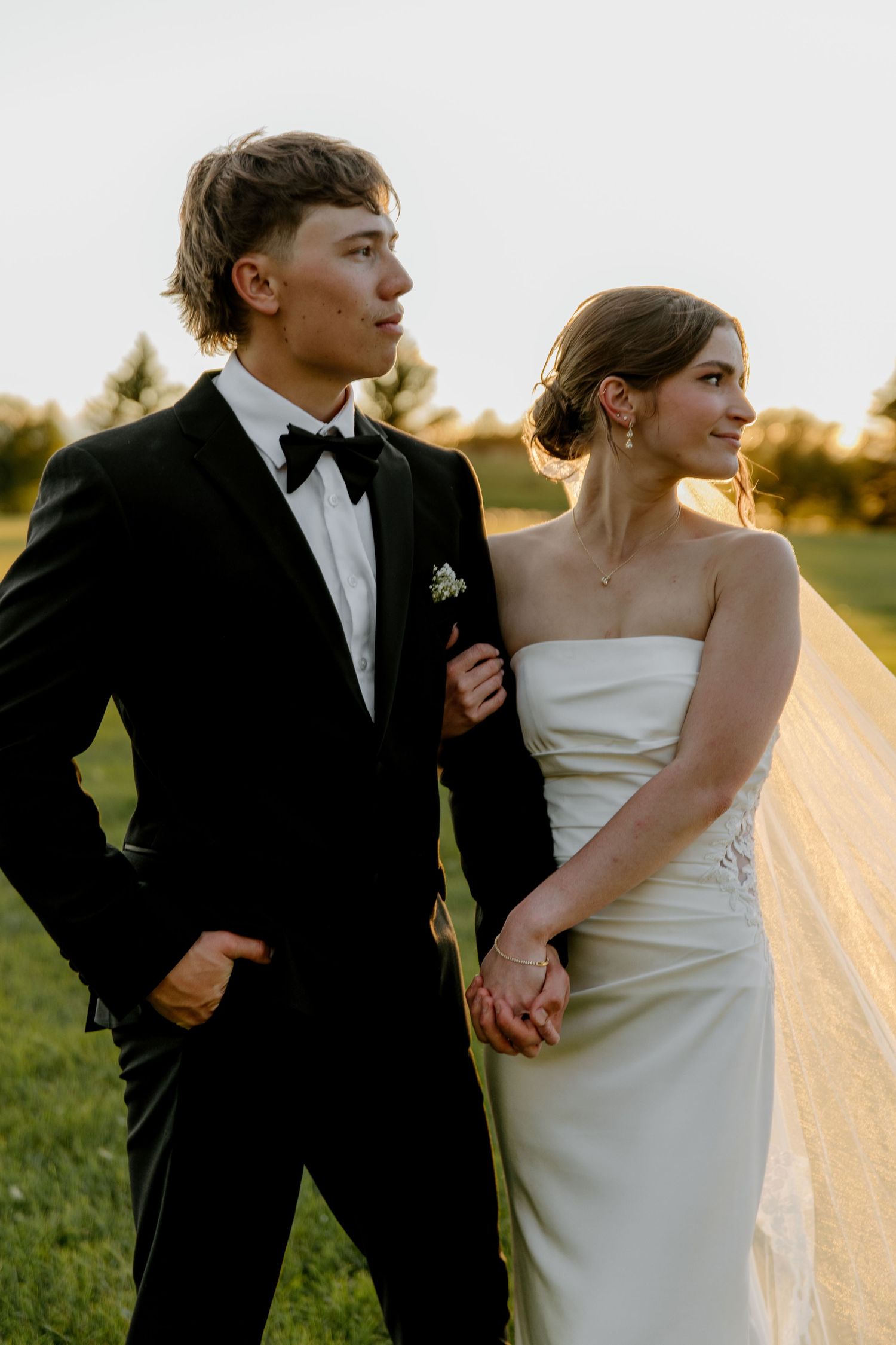 A young couple in wedding attire, groom in black tuxedo and bride in white strapless gown, posing outdoors at sunset.