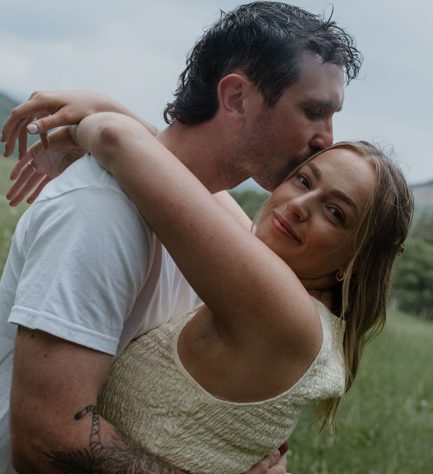 Romantic couple embracing in a rainy outdoor setting, man kissing womans head while she smiles softly.