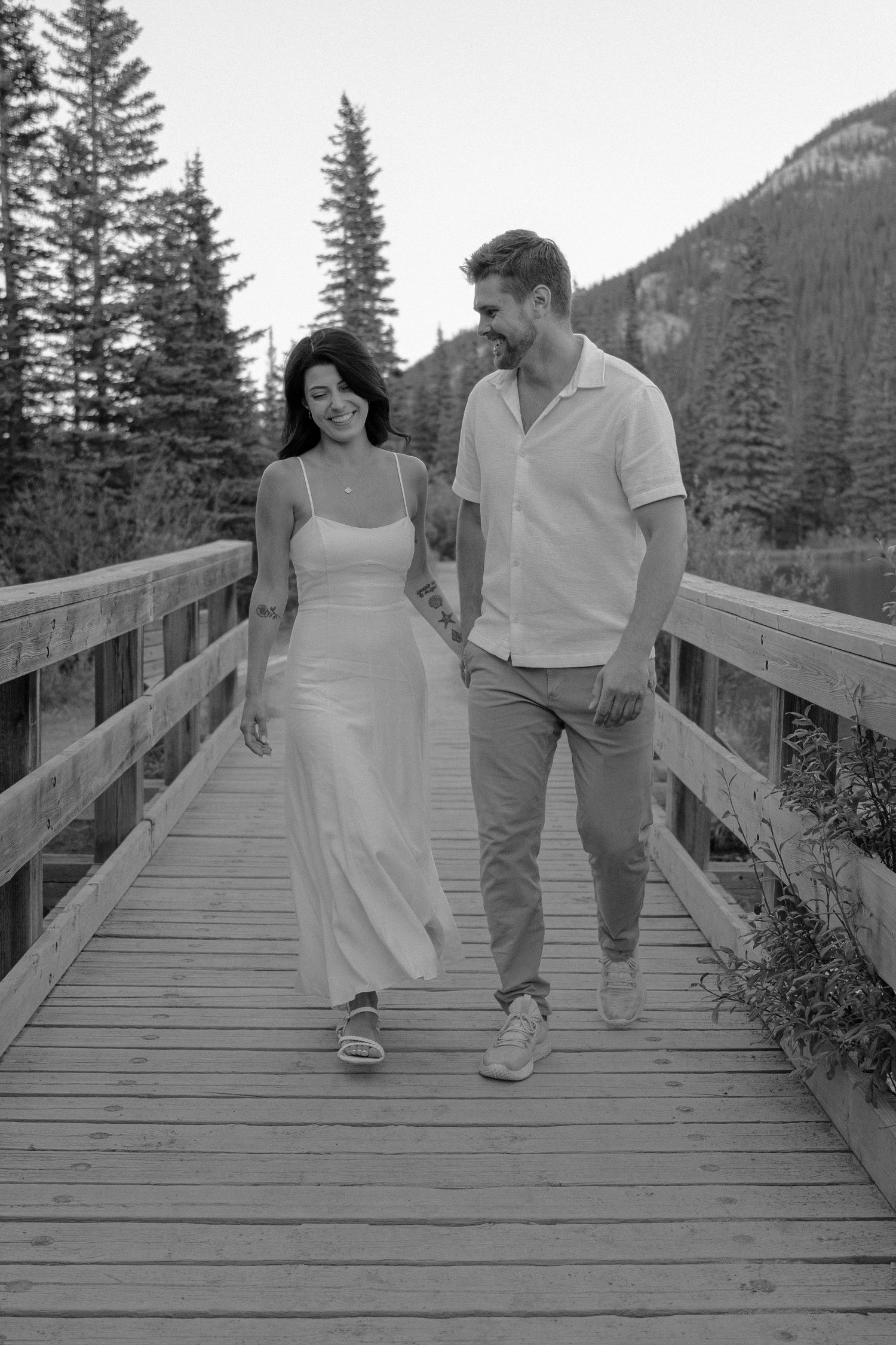 Black and white photo of a couple holding hands and walking on a wooden bridge surrounded by pine trees and mountains.