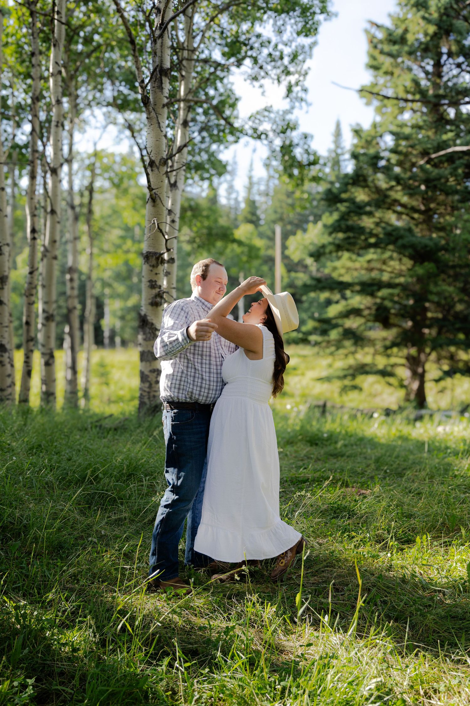 Couple dancing outdoors in a lush green forest with aspen trees, woman in white dress and hat, man in plaid shirt.