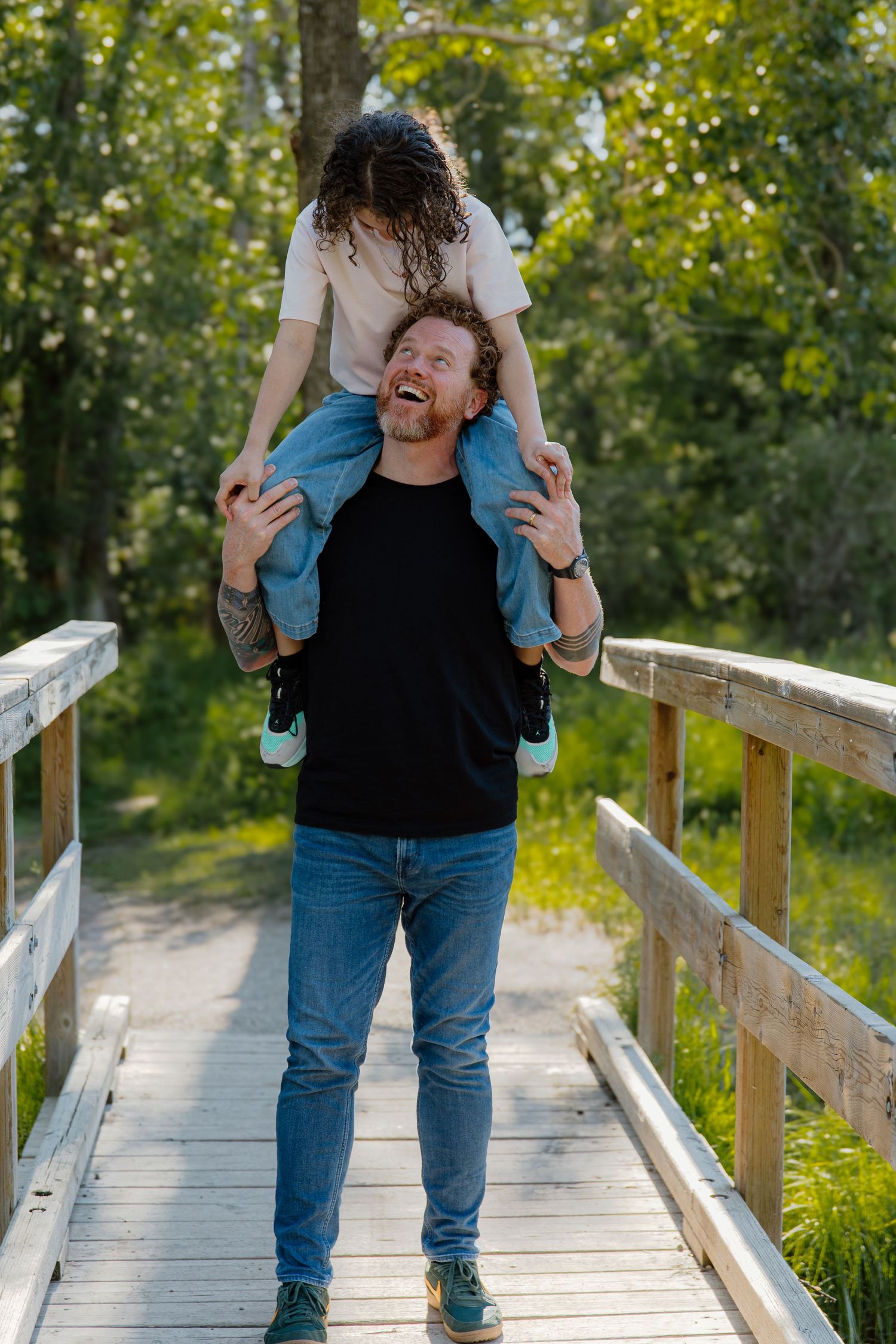 A laughing man carries a child on his shoulders on a wooden bridge surrounded by lush green trees.