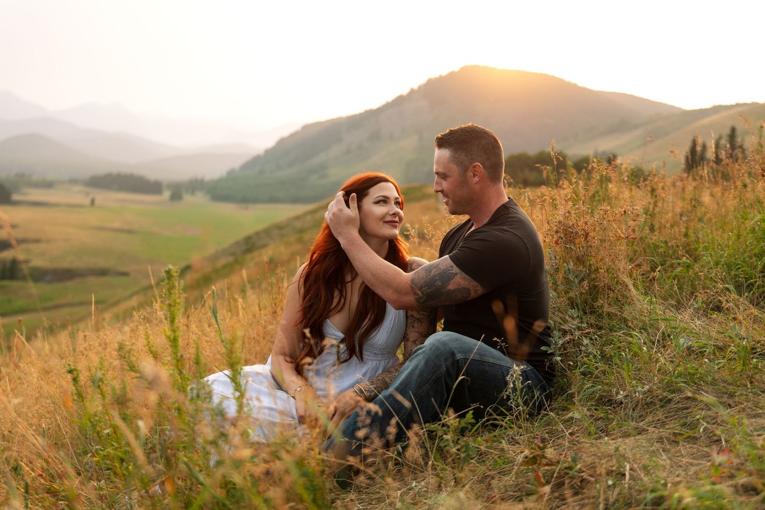 Couple sitting in golden grass on a hillside at sunset with mountain valley views in the background.