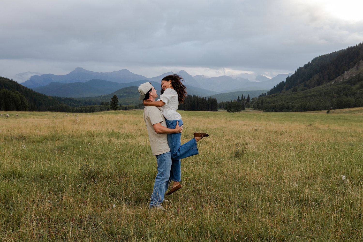 Couple embracing in a green mountain meadow under cloudy skies, man lifting woman in a romantic outdoor setting.