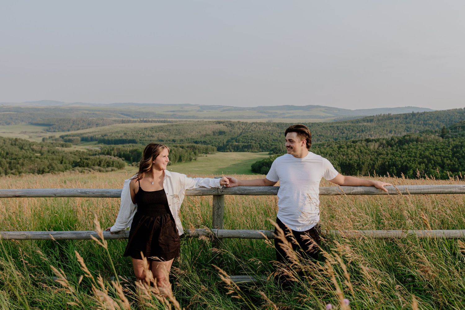 Couple holding hands at a wooden fence overlooking a lush green valley landscape on a sunny day.