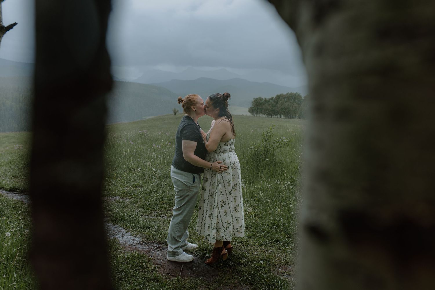 Couple sharing a romantic kiss on a misty green hillside, framed by trees in a dreamy outdoor engagement photo.