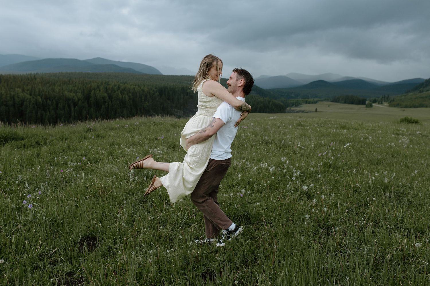Couple embracing in a green mountain meadow, man lifting woman in white dress under cloudy skies.