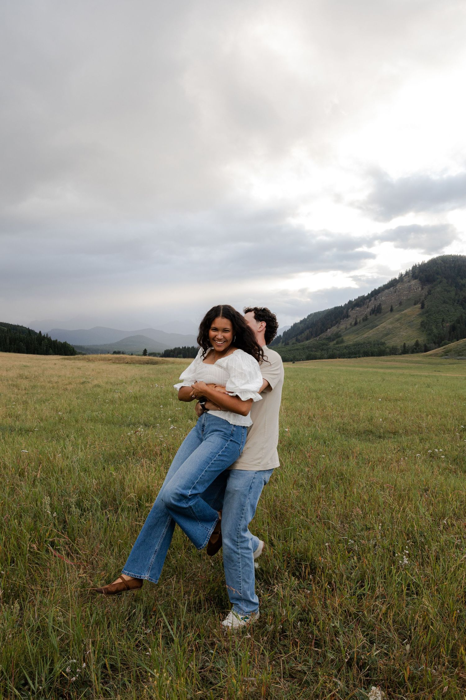 Couple embracing and dancing in a green meadow with mountain views under a cloudy sky, both wearing jeans and white tops.