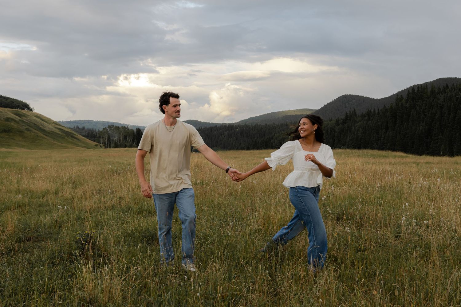 A couple holds hands and walks through a lush green meadow with mountains and dramatic clouds in the background.