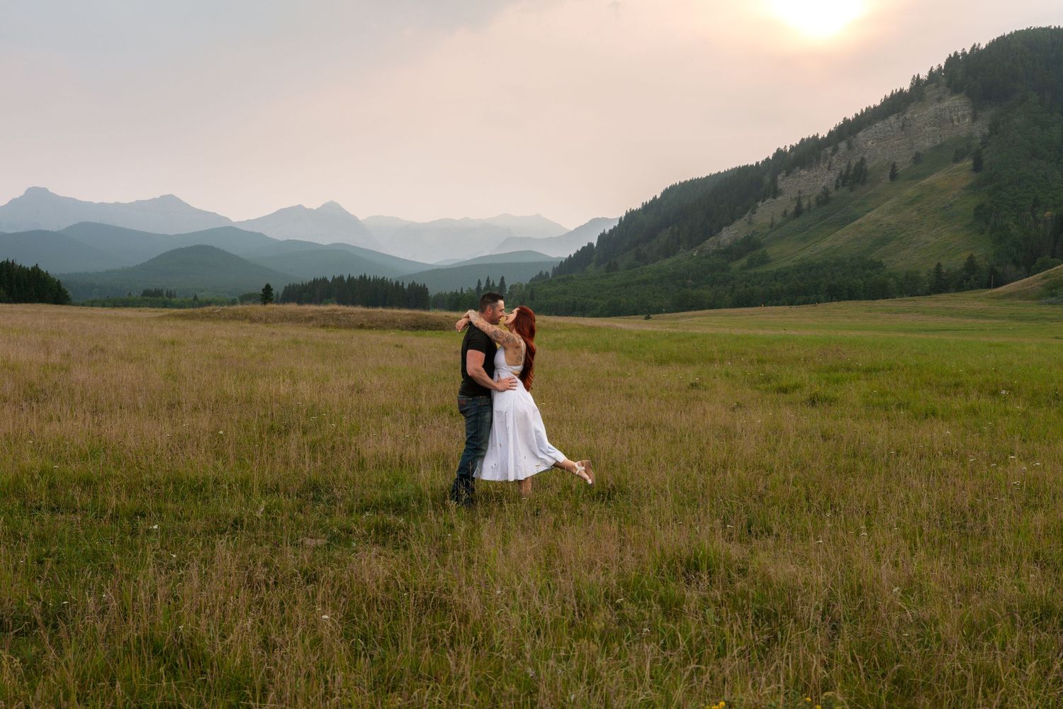 Couple sharing a romantic kiss in a vast mountain meadow at sunset, bride in white dress, scenic peaks in background.