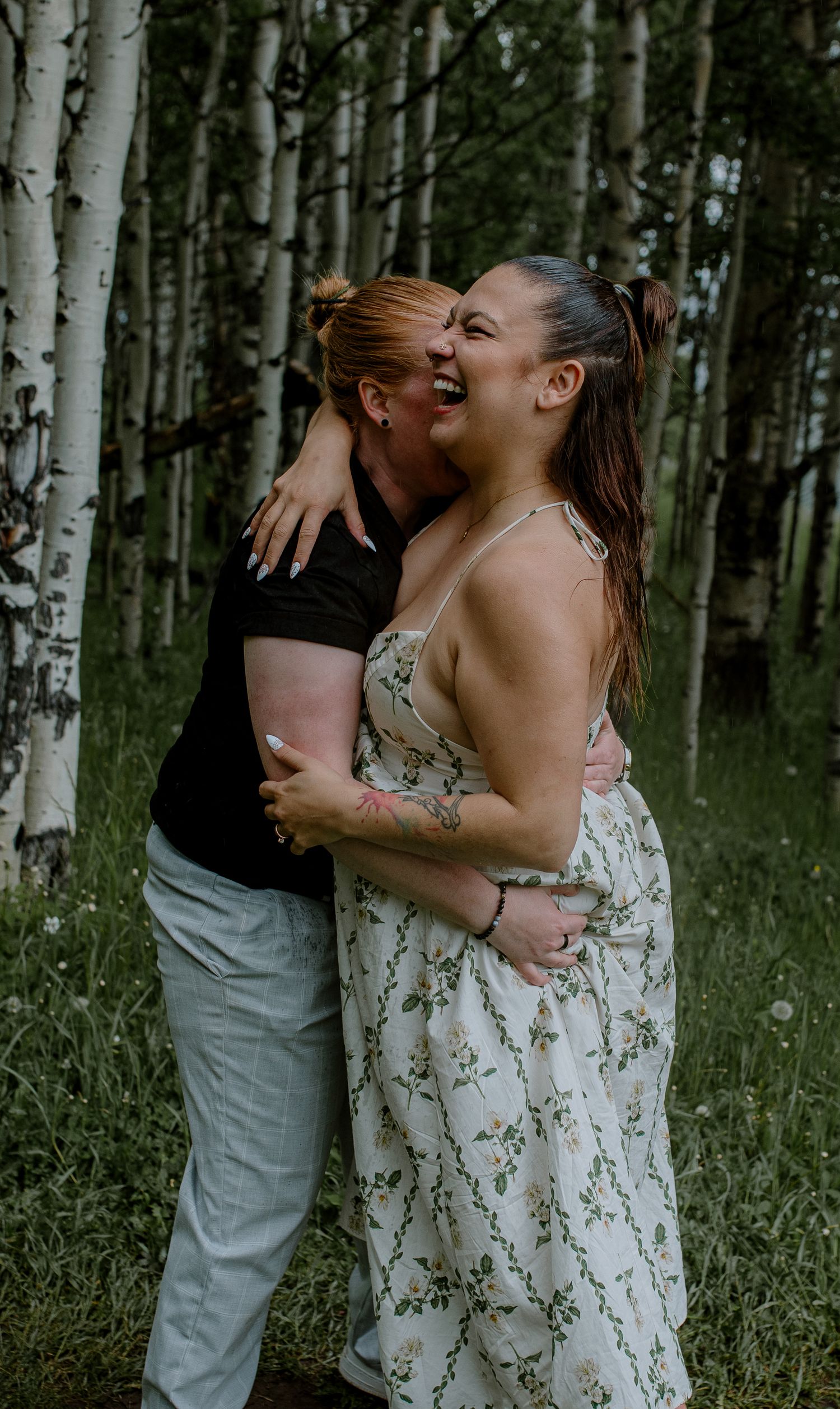 Two women embracing and laughing together among white birch trees in a sunny forest setting.