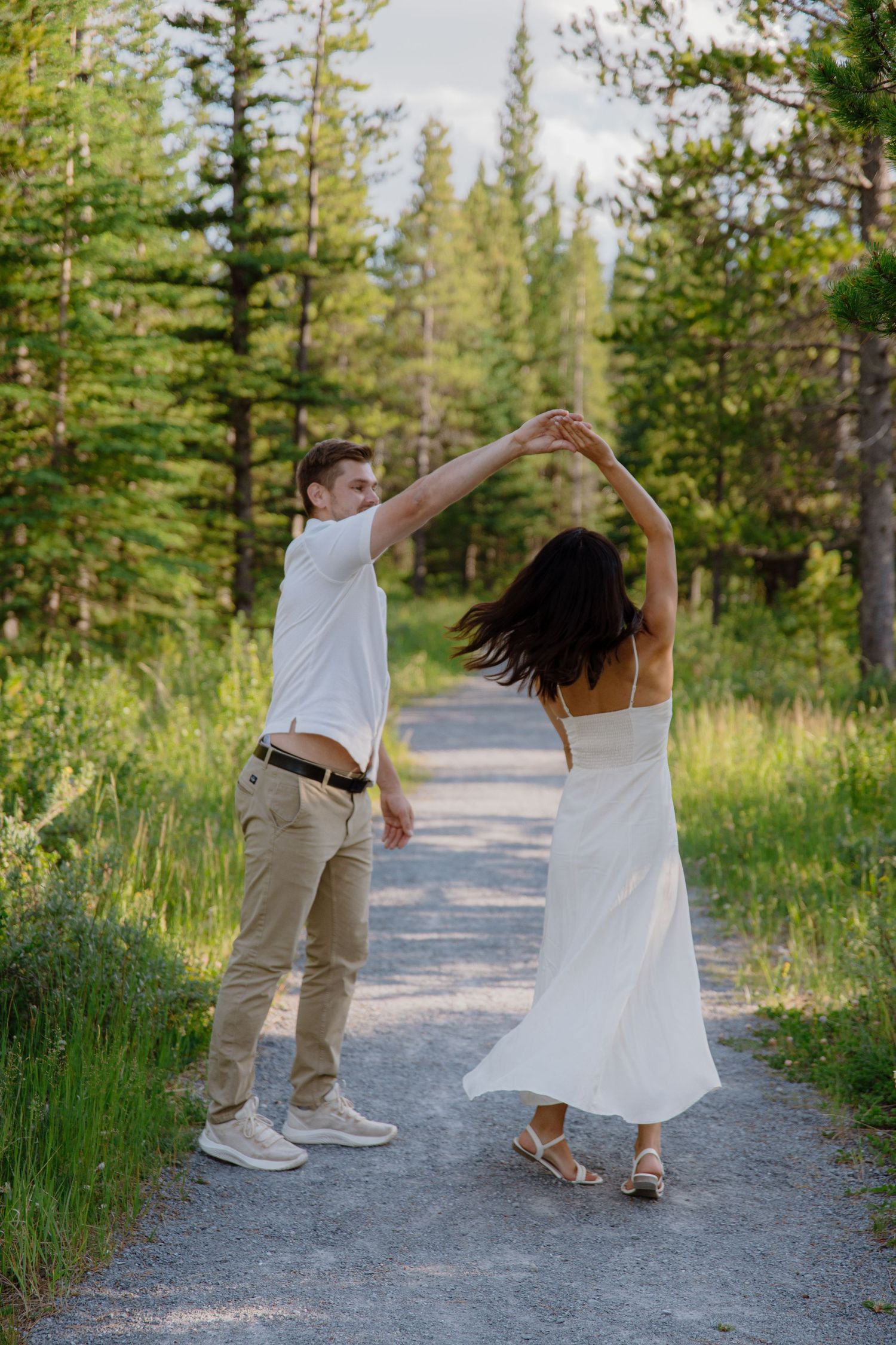 A couple dances on a forest path, man in white shirt spinning woman in flowing white dress among tall green trees.