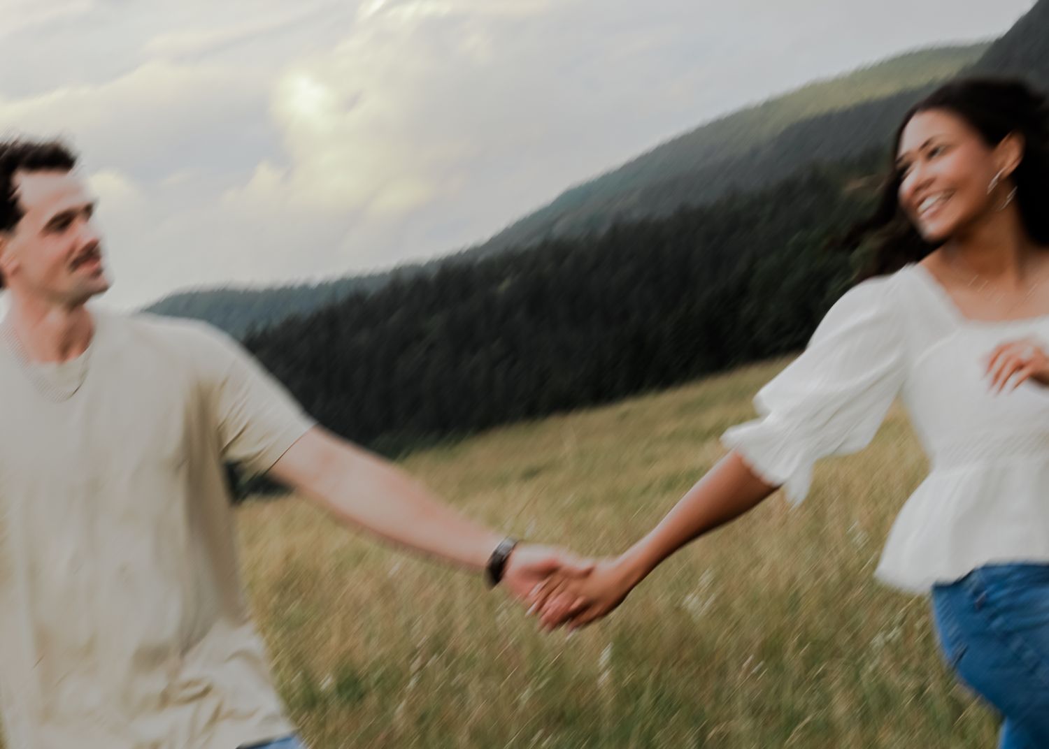 A joyful couple holds hands running through a lush green meadow with misty mountains in the background.