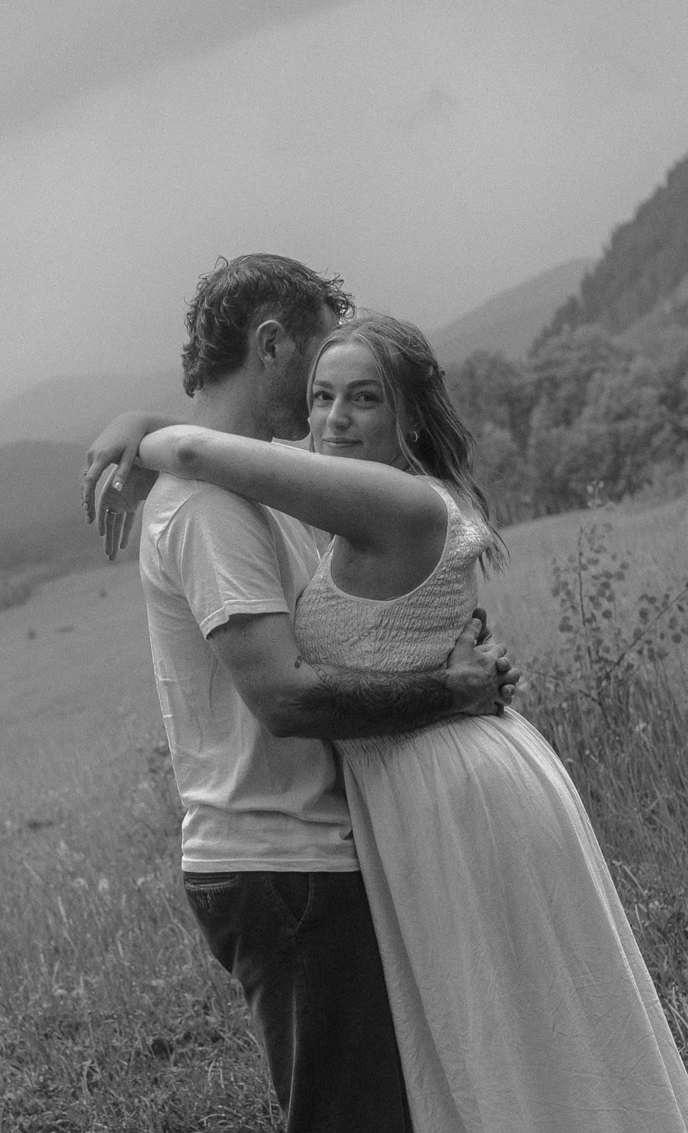 Black and white photo of a couple embracing in a mountain valley landscape.