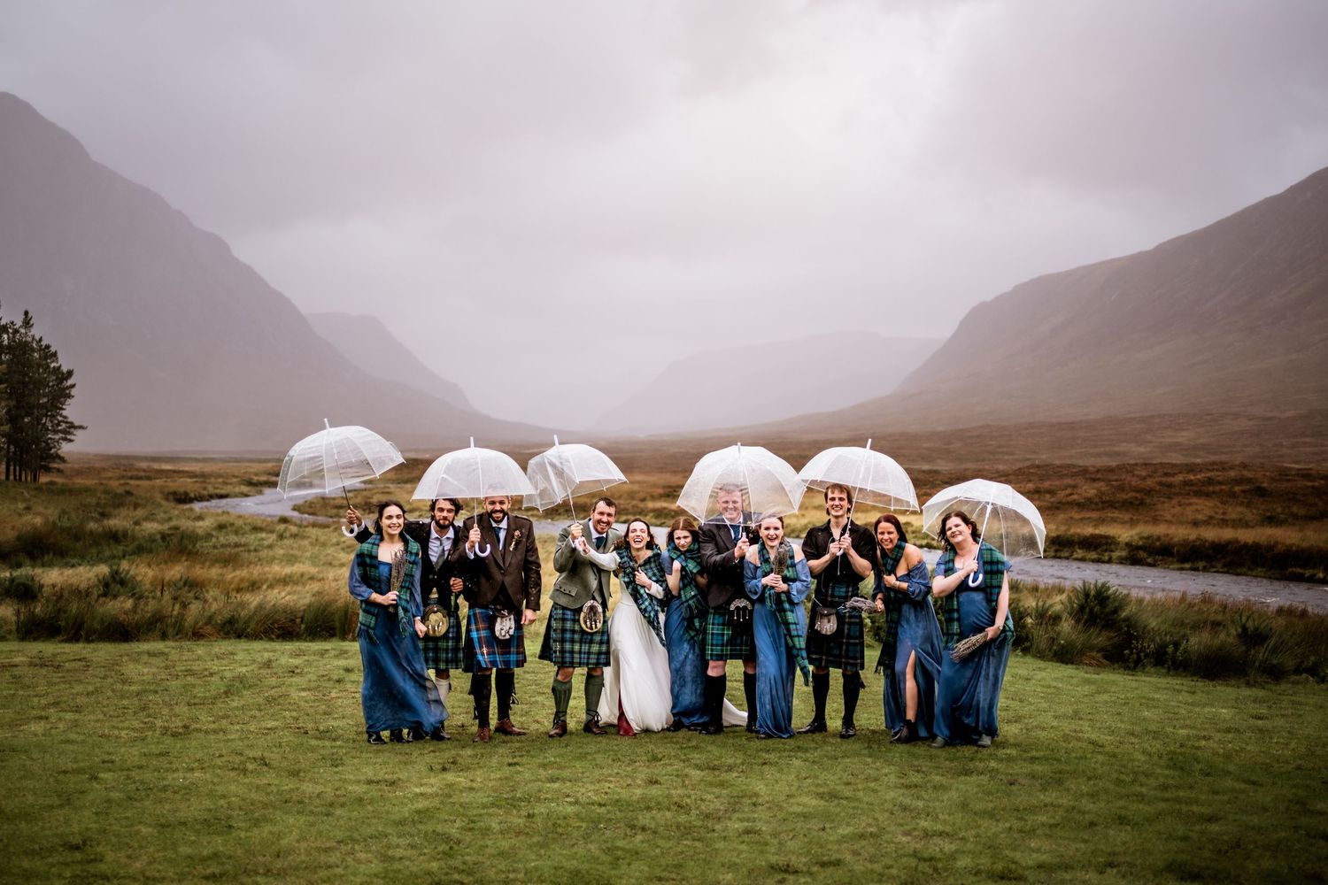 A wedding party poses with white umbrellas against a misty Scottish highland backdrop. Kingshouse Hotel Wedding Photography