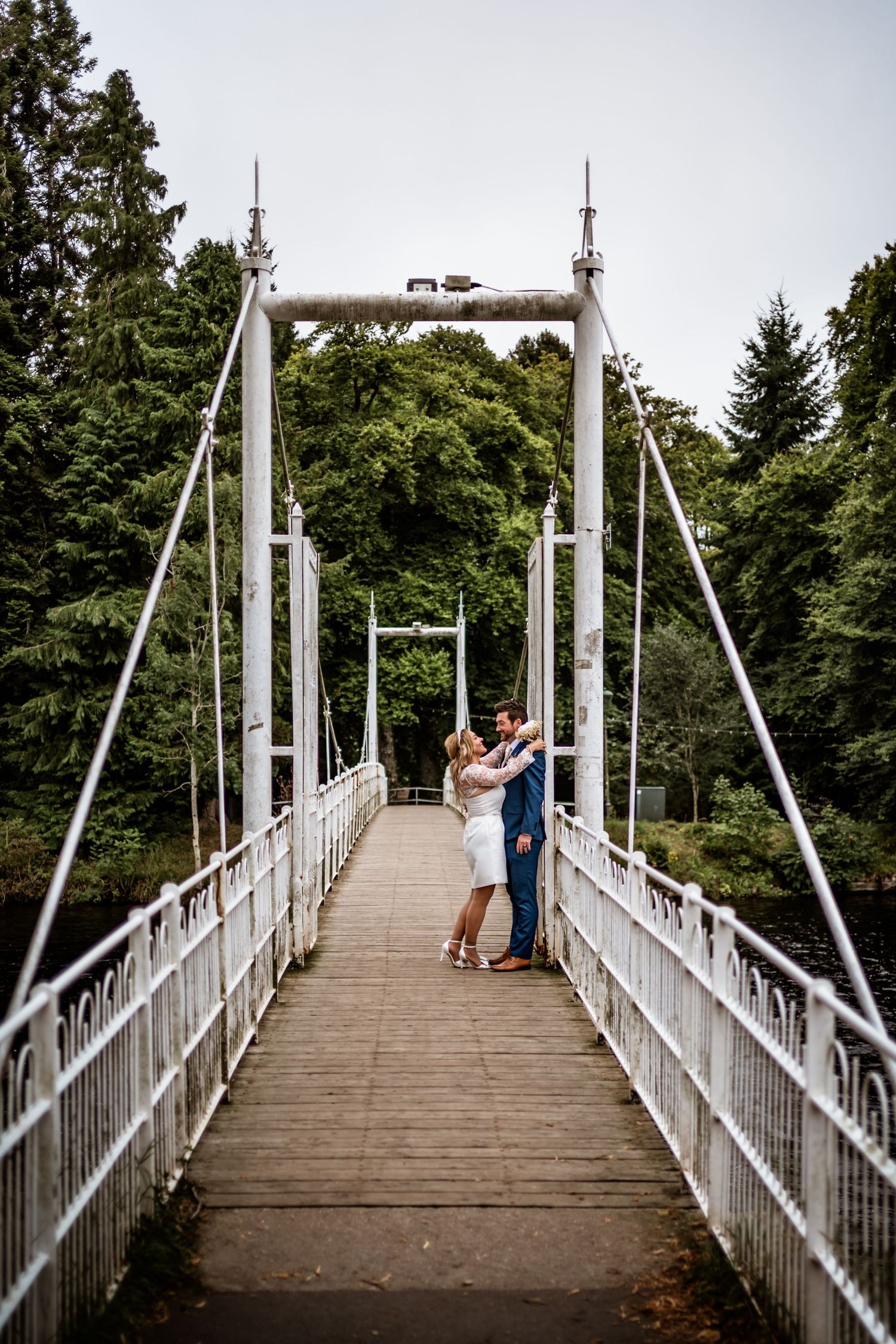 A couple sharing an intimate moment on a white suspension bridge surrounded by trees. Scottish Highlands Inverness Wedding Photography