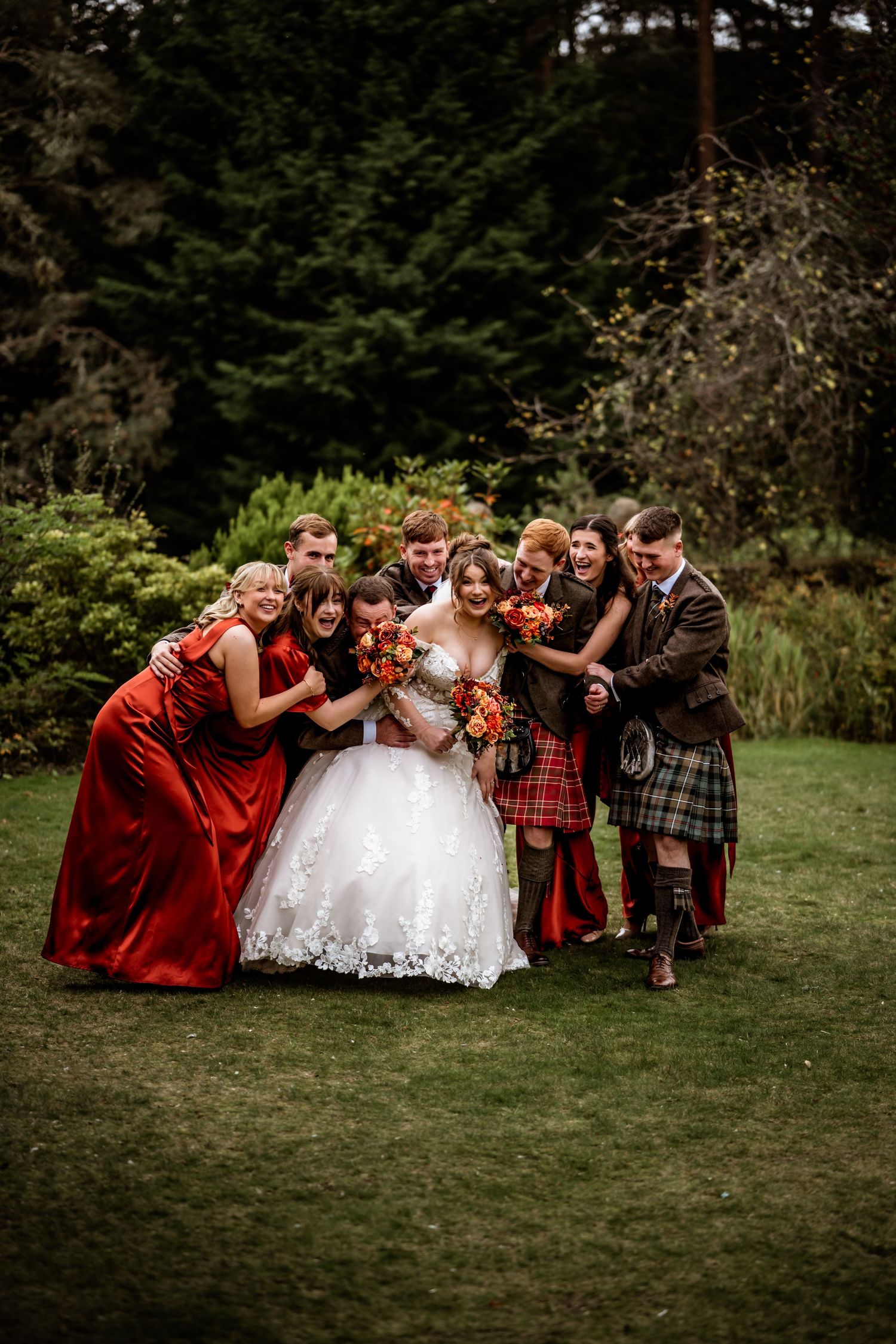 Wedding group photo with bridesmaids in red dresses and groomsmen in kilts on grass. Loch Ness Country House Hotel Inverness Wedding