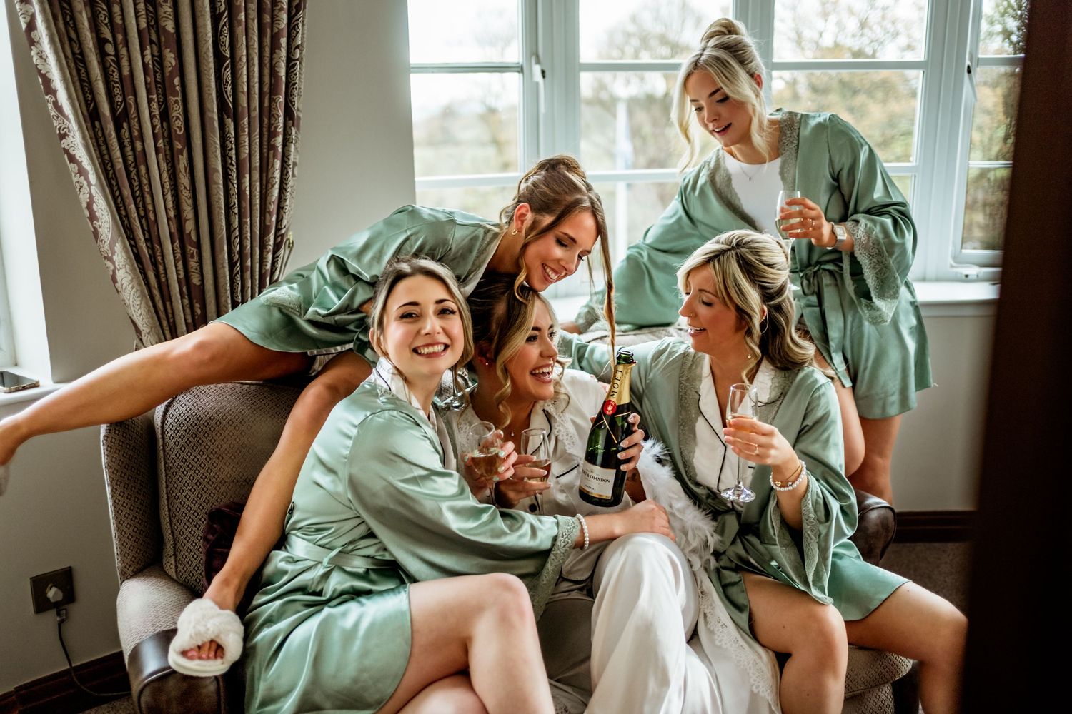A group wearing matching mint green silk robes lounges together on a couch during wedding preparations.