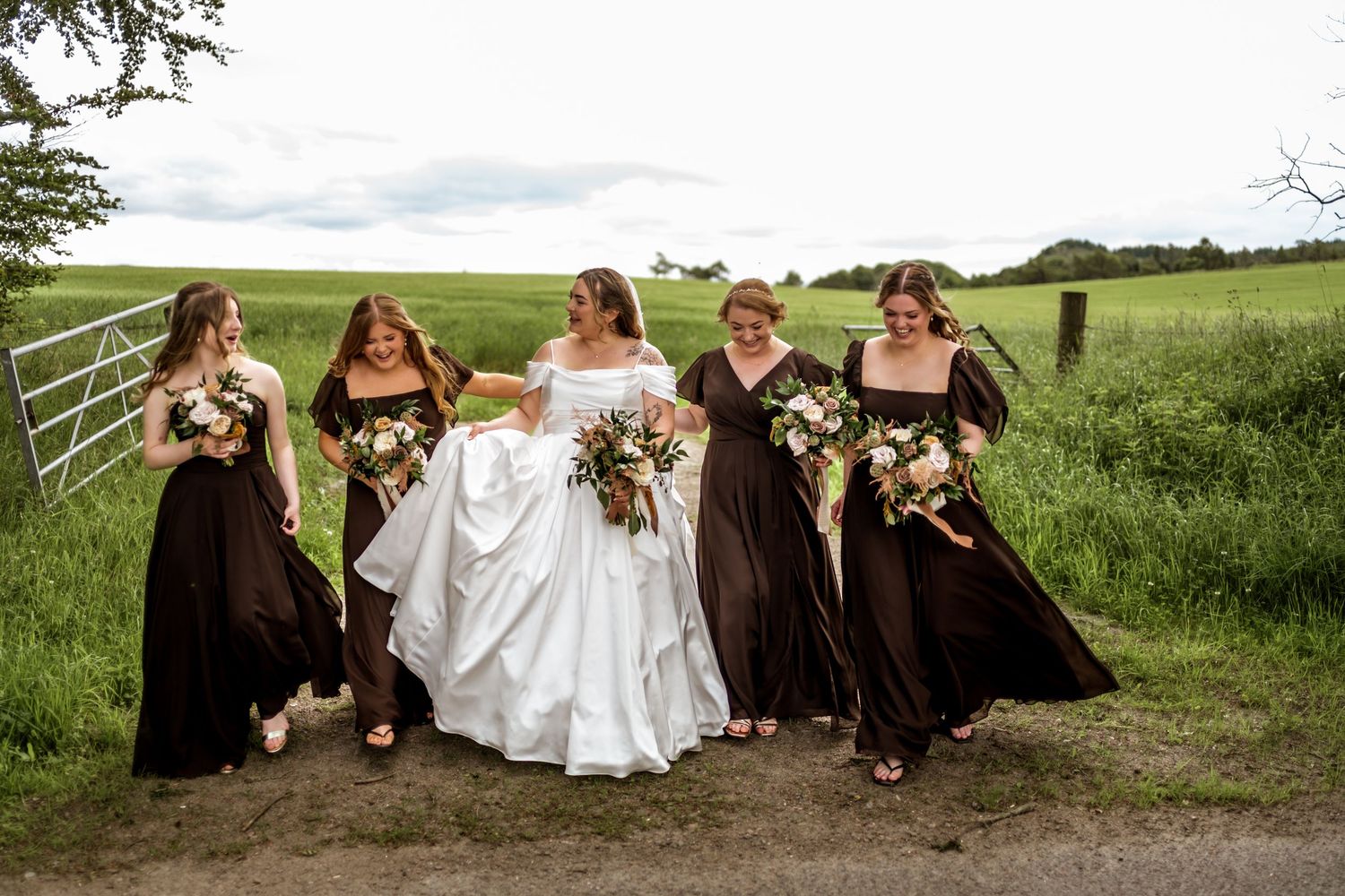 Bride in white gown walks with four bridesmaids in dark brown dresses, holding bouquets on a scenic countryside path.
