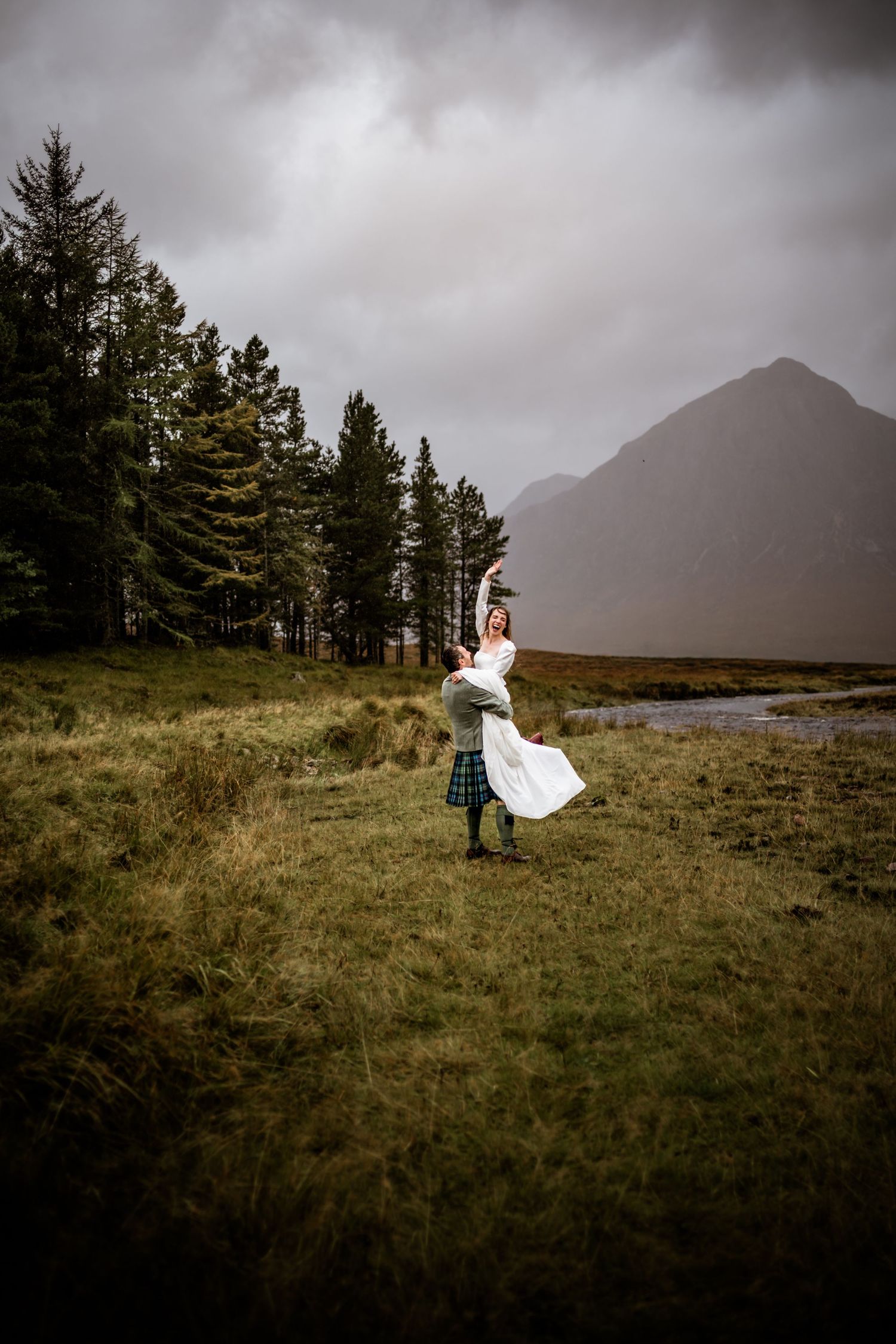 A white wedding dress flows in the wind against a dramatic mountain landscape with evergreen trees. Kingshouse Hotel Wedding Photography