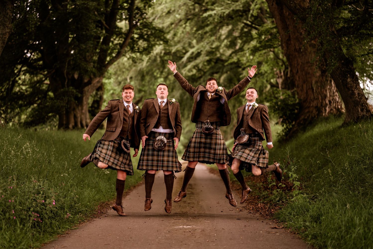 Four young men in traditional Scottish kilts and jackets jumping joyfully on a tree-lined path.