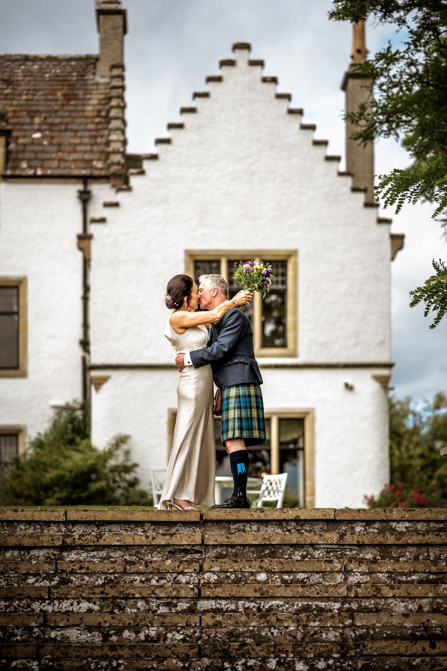 A couple embracing in front of a traditional Scottish white house with stepped gables. Scottish Highlands Wedding Photography