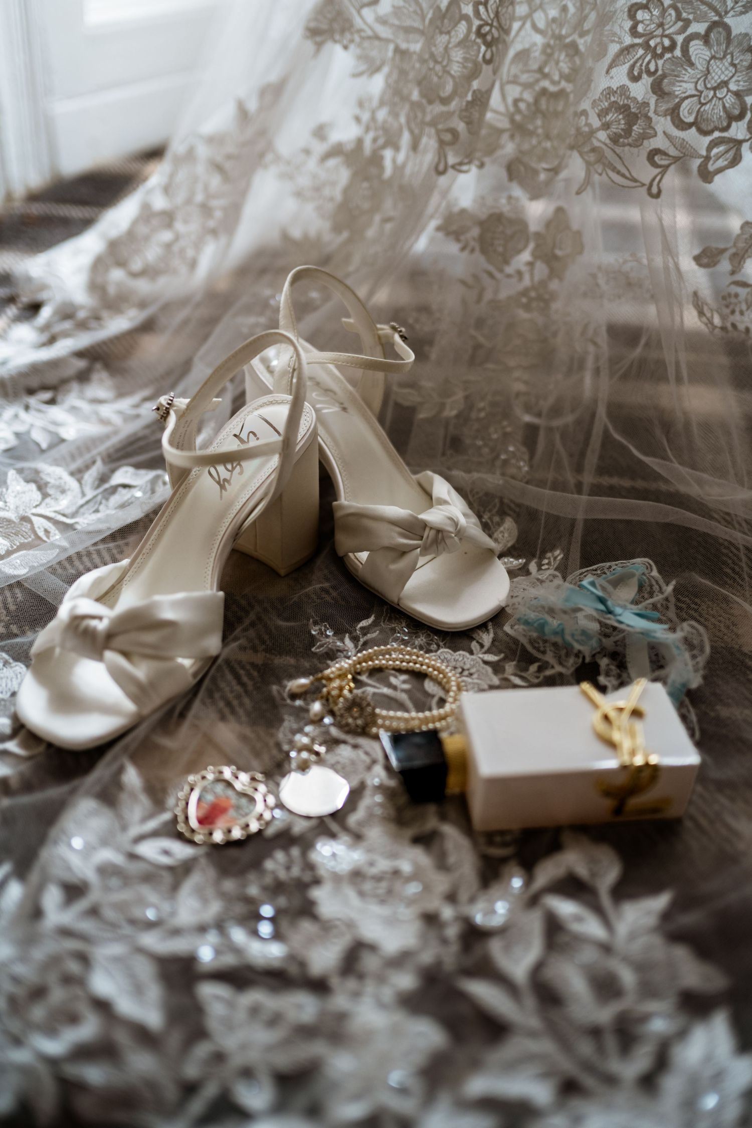 Bridal flat lay featuring ivory block heel sandals, gold jewelry, YSL perfume, and lace wedding veil on wooden floor.