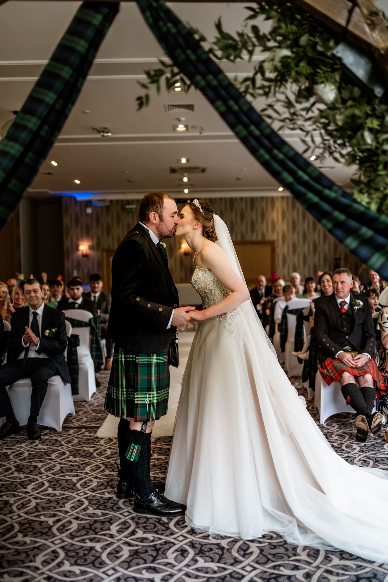 Bride and groom share first kiss in Scottish wedding ceremony, groom wearing green tartan kilt, guests seated in elegant ballroom.
