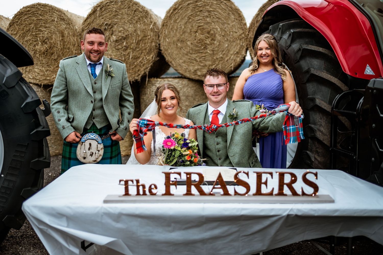 A wedding party poses with a tractor and hay bales featuring a personalized name sign.