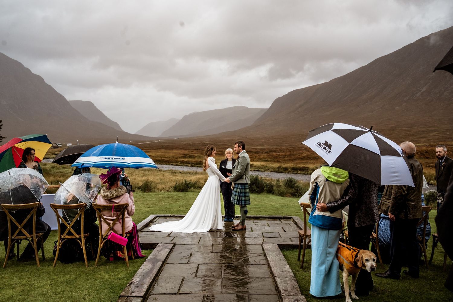 An outdoor wedding ceremony in the Scottish highlands with guests holding umbrellas under cloudy skies. Kingshouse Hotel Wedding Photography