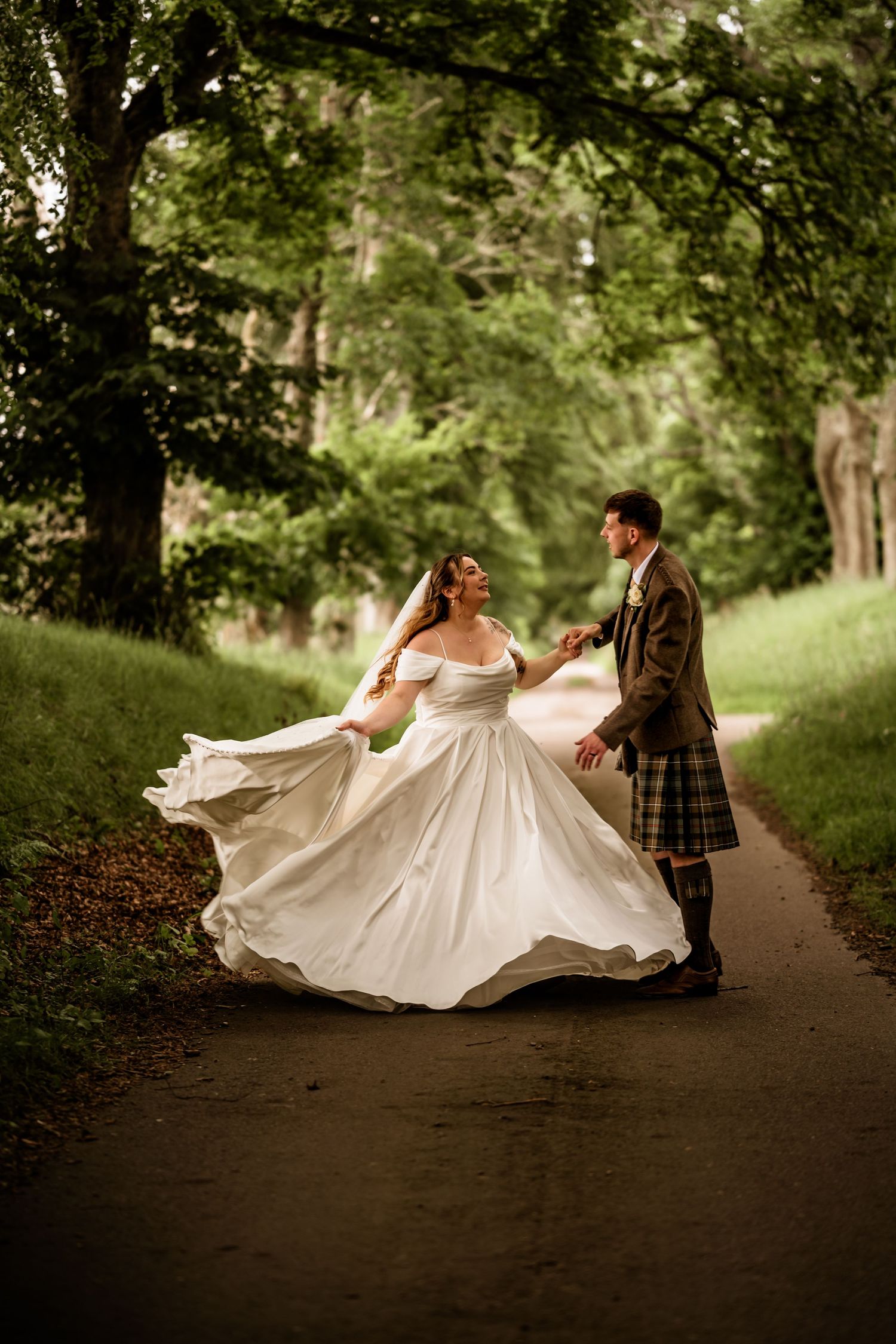 Bride in flowing white gown twirls with groom in tartan kilt on a tree-lined path during outdoor wedding.