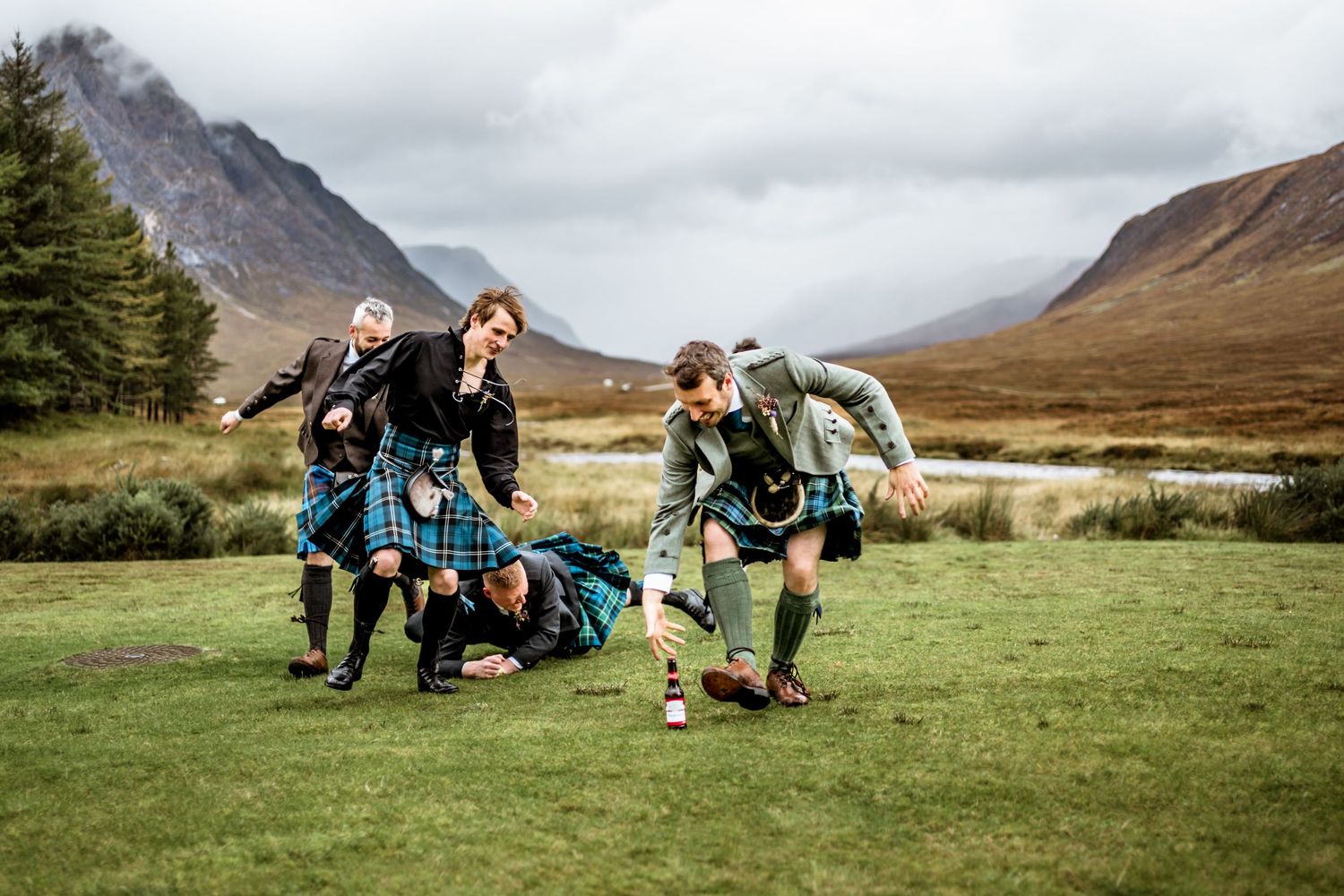 Wedding party in kilts playfully poses in the Scottish Highlands with mountains in background. Kingshouse Hotel Wedding Photography
