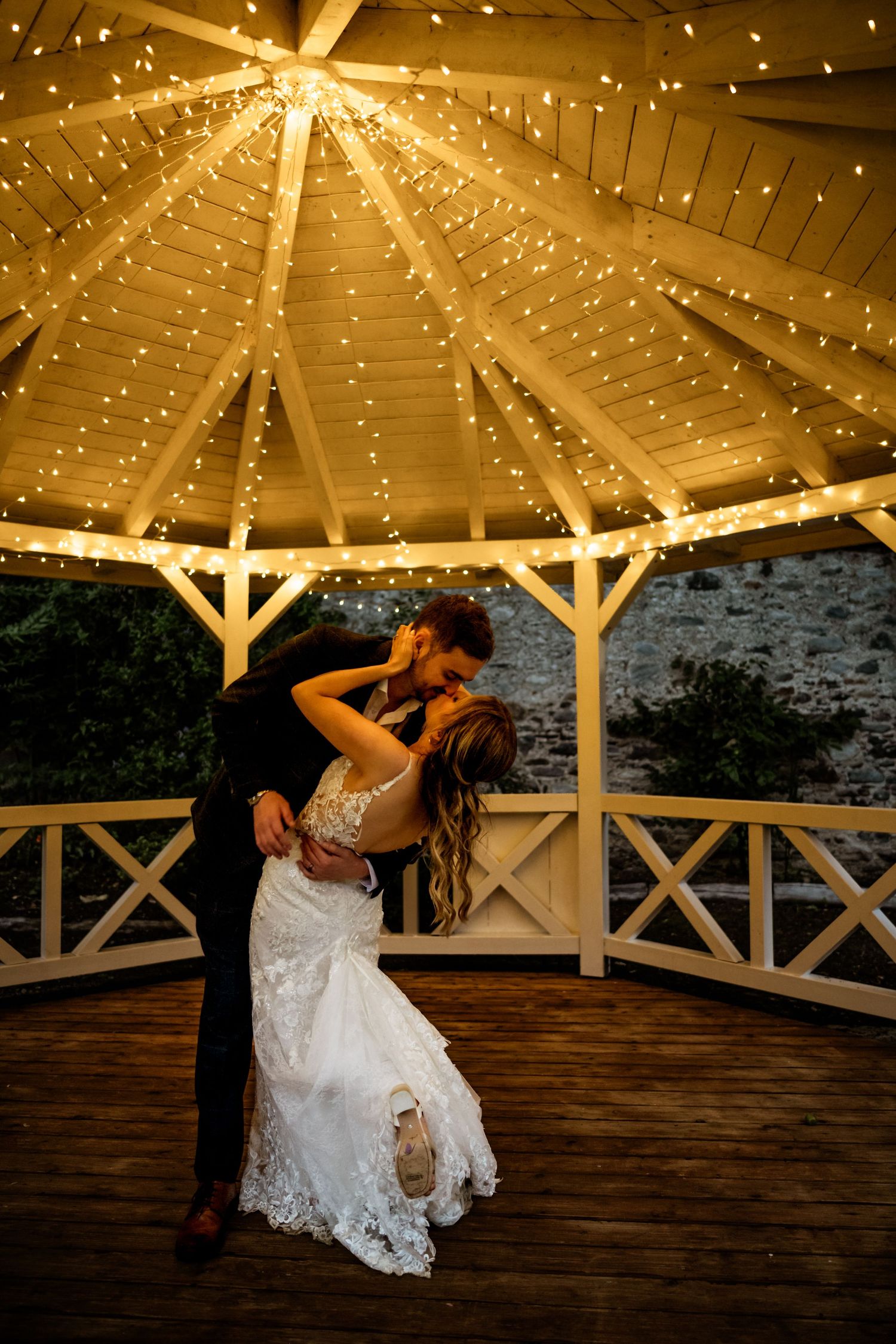 Bride and groom share a romantic kiss under a glowing gazebo adorned with fairy lights at their wedding reception.