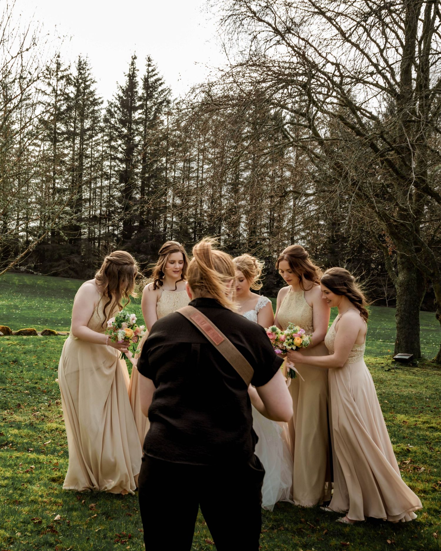 Wedding party in cream colored dresses gathered in circle outdoors among trees.