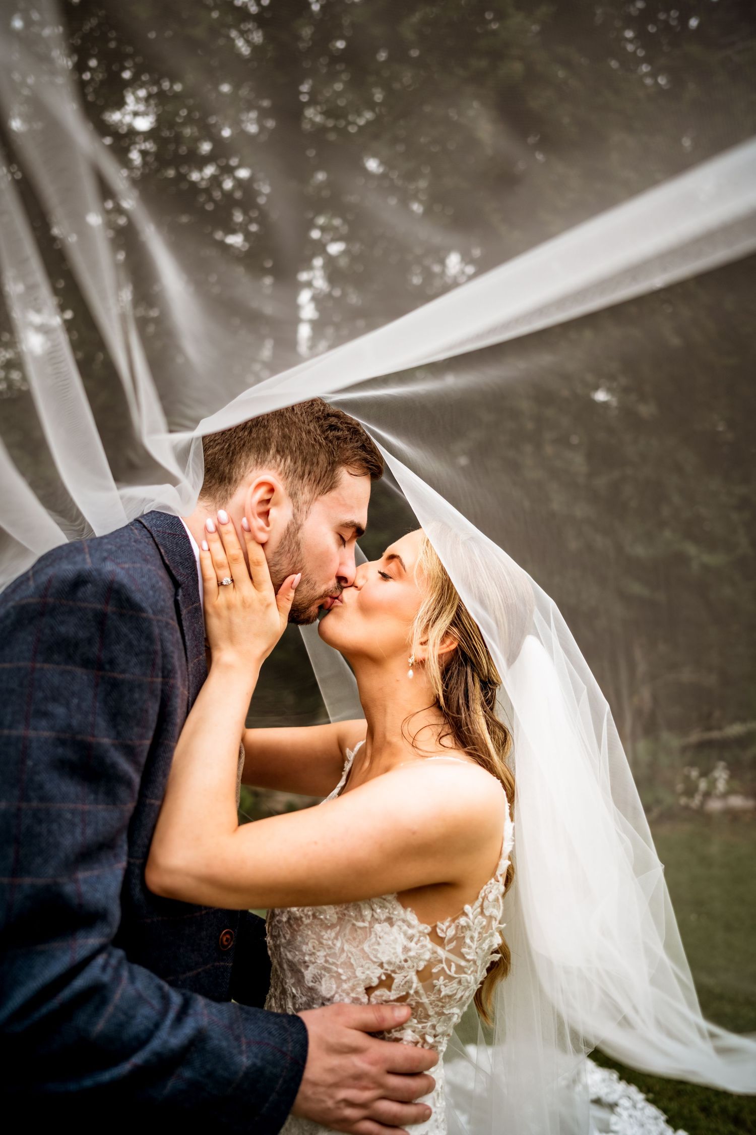 Bride and groom share a romantic kiss under a flowing white veil outdoors on their wedding day.