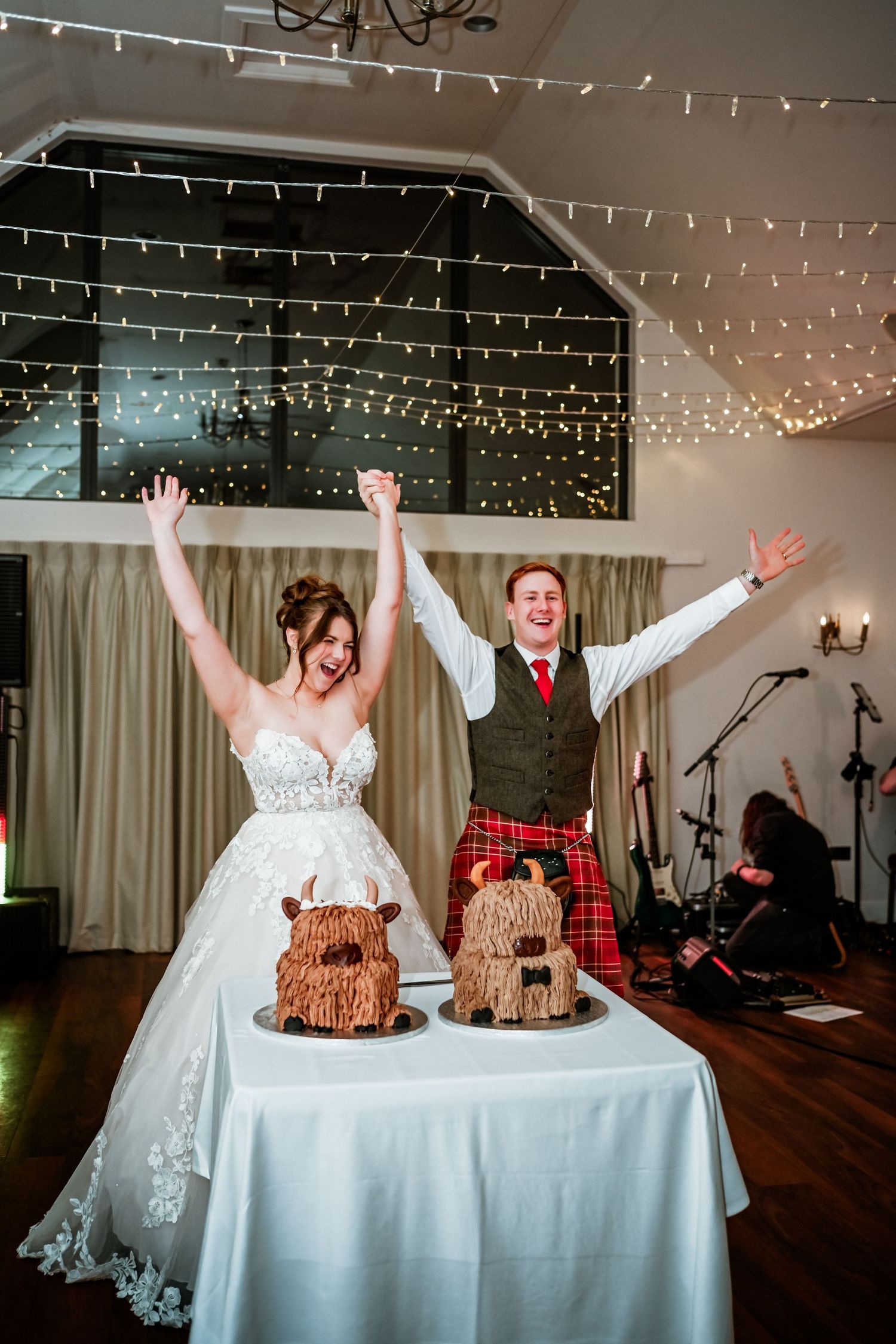 Wedding couple celebrates behind their cake table with fairy lights twinkling overhead. LochNess Country House Hotel Wedding Photography
