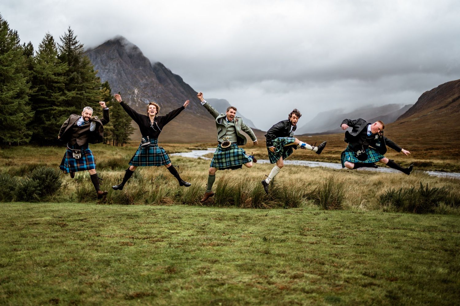 People in traditional Scottish kilts leaping joyfully with mountains and cloudy skies in background. Kingshouse Hotel Wedding Photography