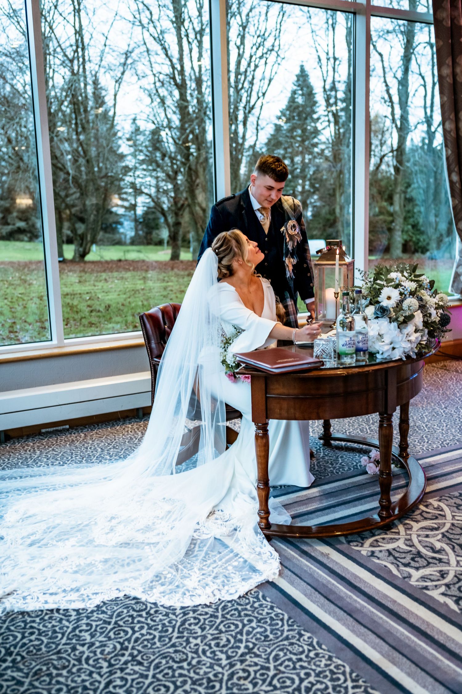 A couple signs their marriage documents at an elegant wooden desk by large windows.