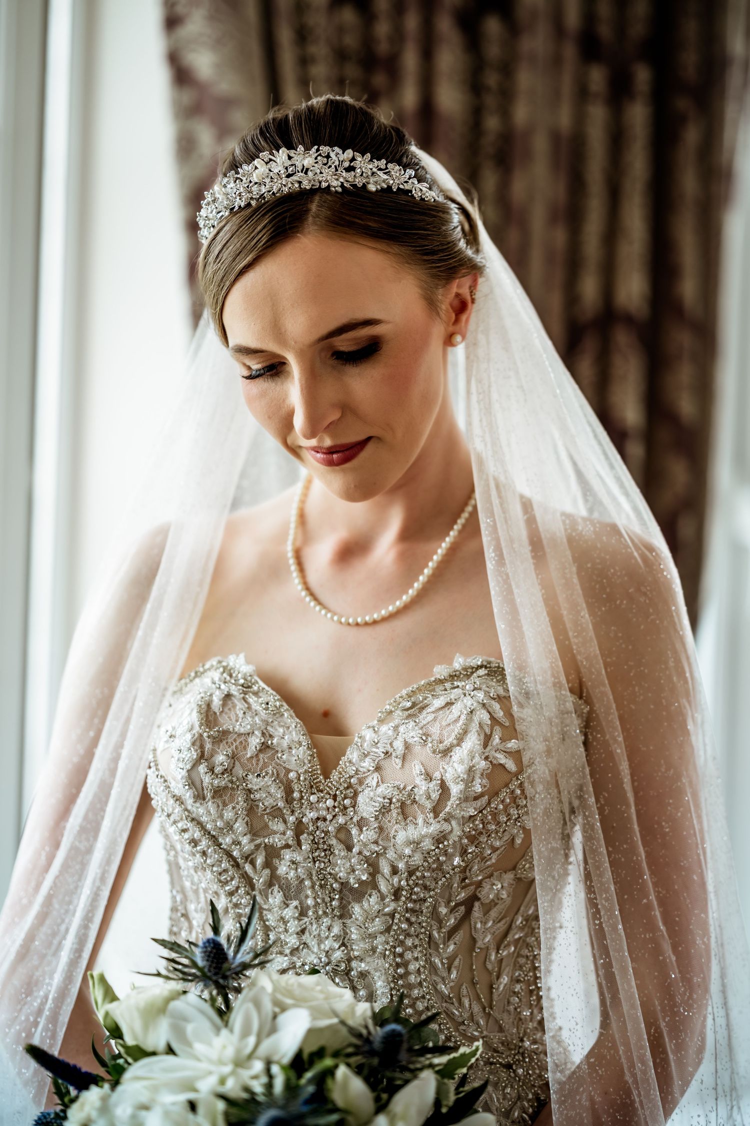 Elegant bride in beaded strapless gown with pearl necklace, crystal tiara, and veil holding white floral bouquet.