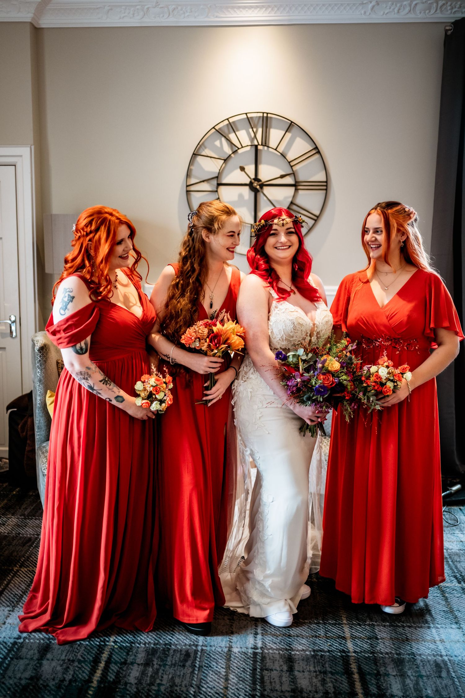 Bridal party in flowing red dresses poses with bride in white under vintage clock. Glen Mhor Hotel Inverness Wedding Photography