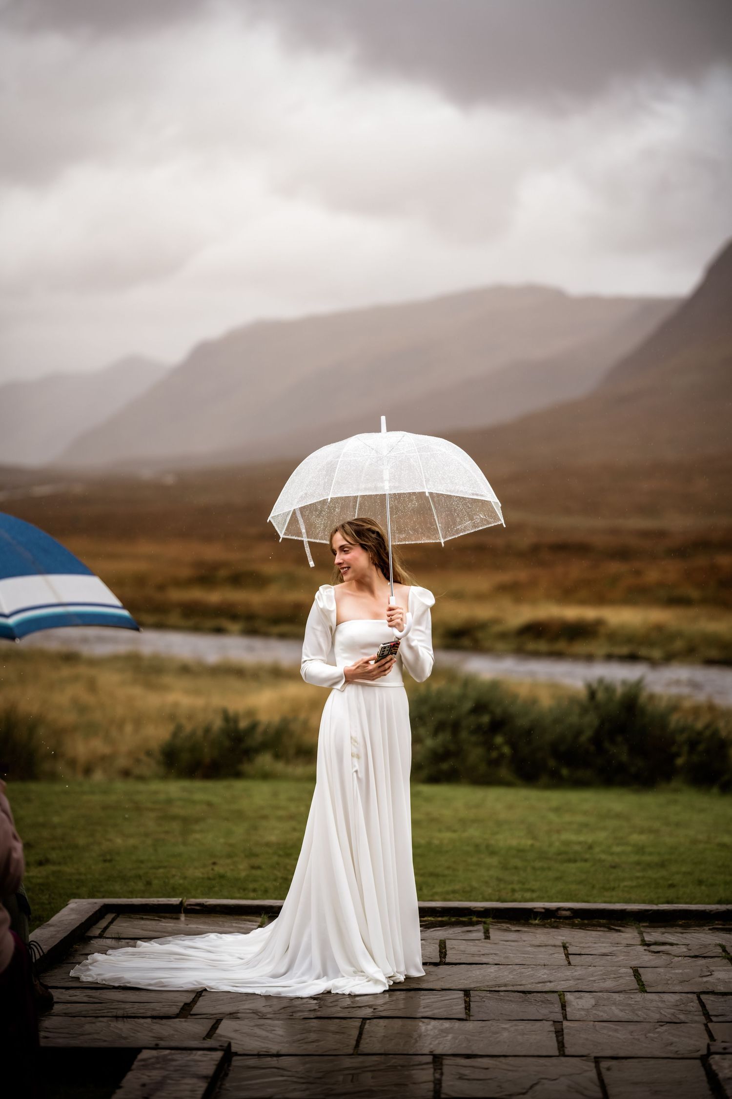 A bride in a white dress holds a clear umbrella against a moody Scottish highland landscape. Kingshouse Hotel Wedding Photography