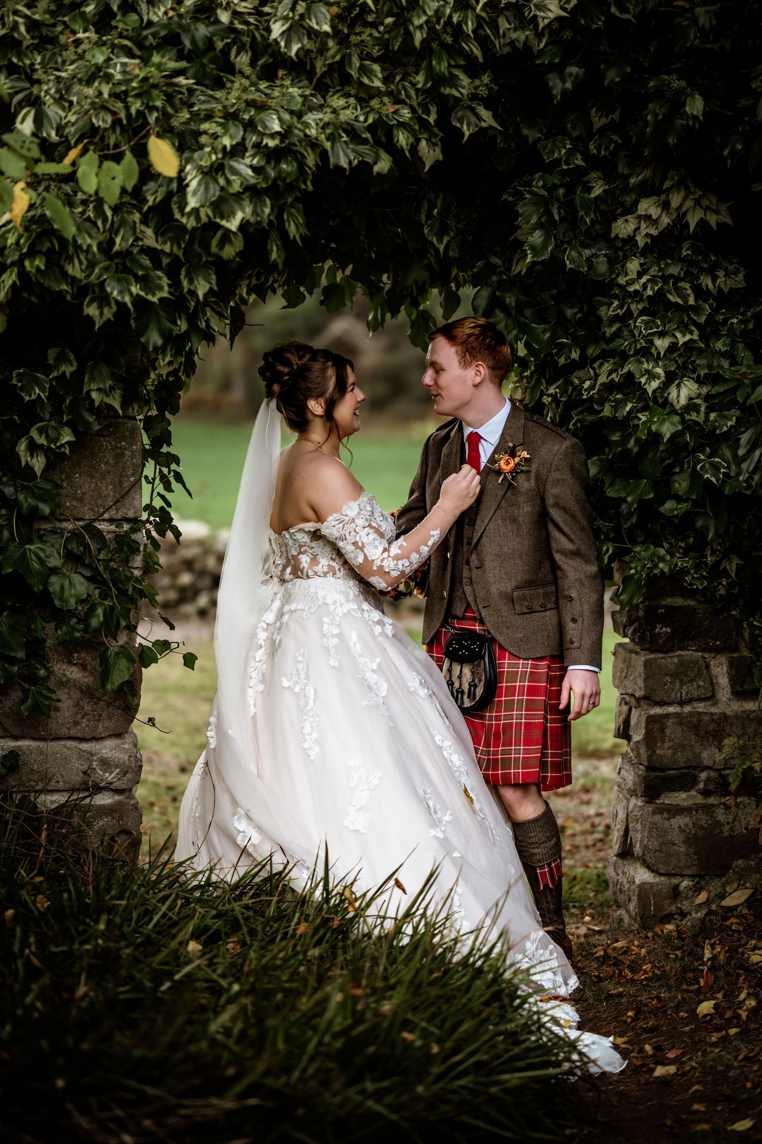 Wedding couple posing among lush green foliage and stone walls in Scottish Highland attire. LochNess Country House Hotel Wedding photography